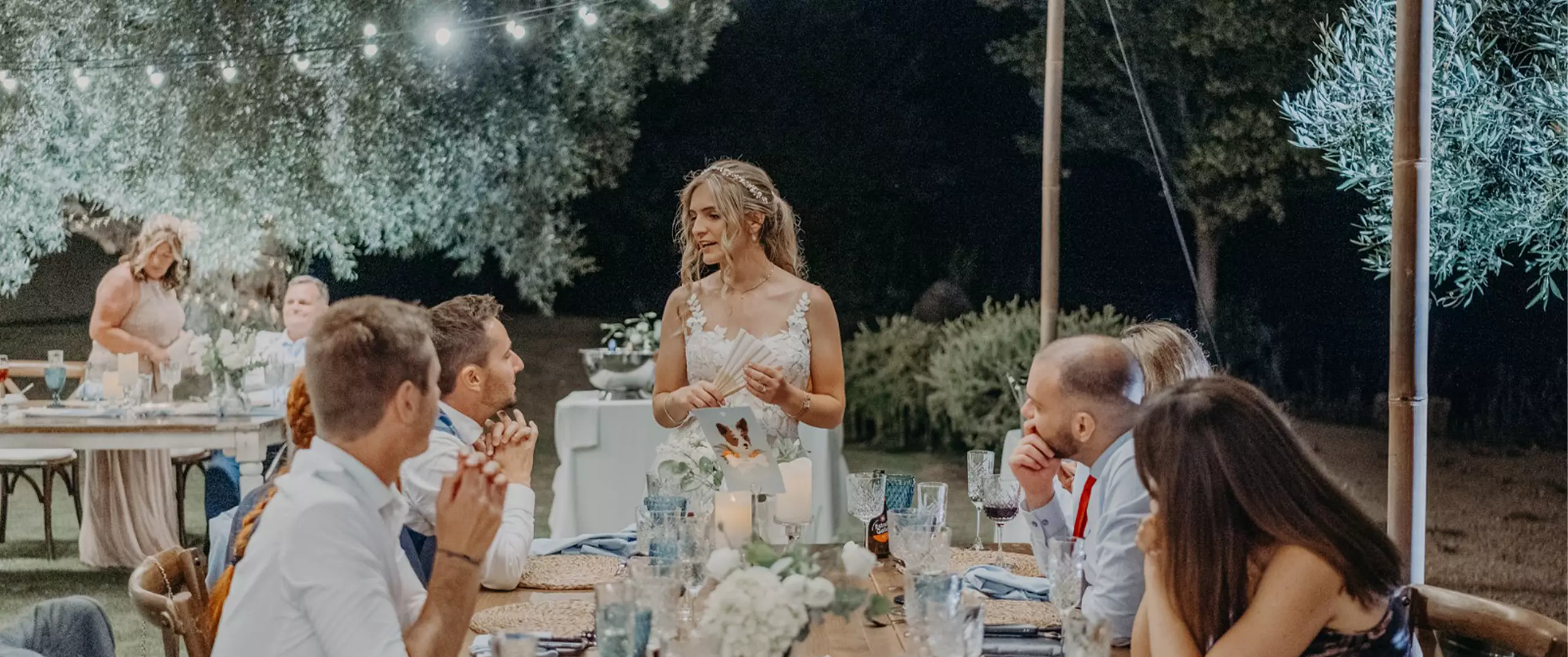 A woman in a white dress stands and speaks to guests seated at a decorated outdoor table during a nighttime event.