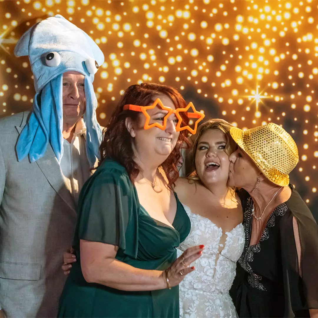 Four people posing for a photo booth; one wears a squid hat, another wears star glasses, a bride smiles, and a woman in a gold hat kisses the bride on the cheek.