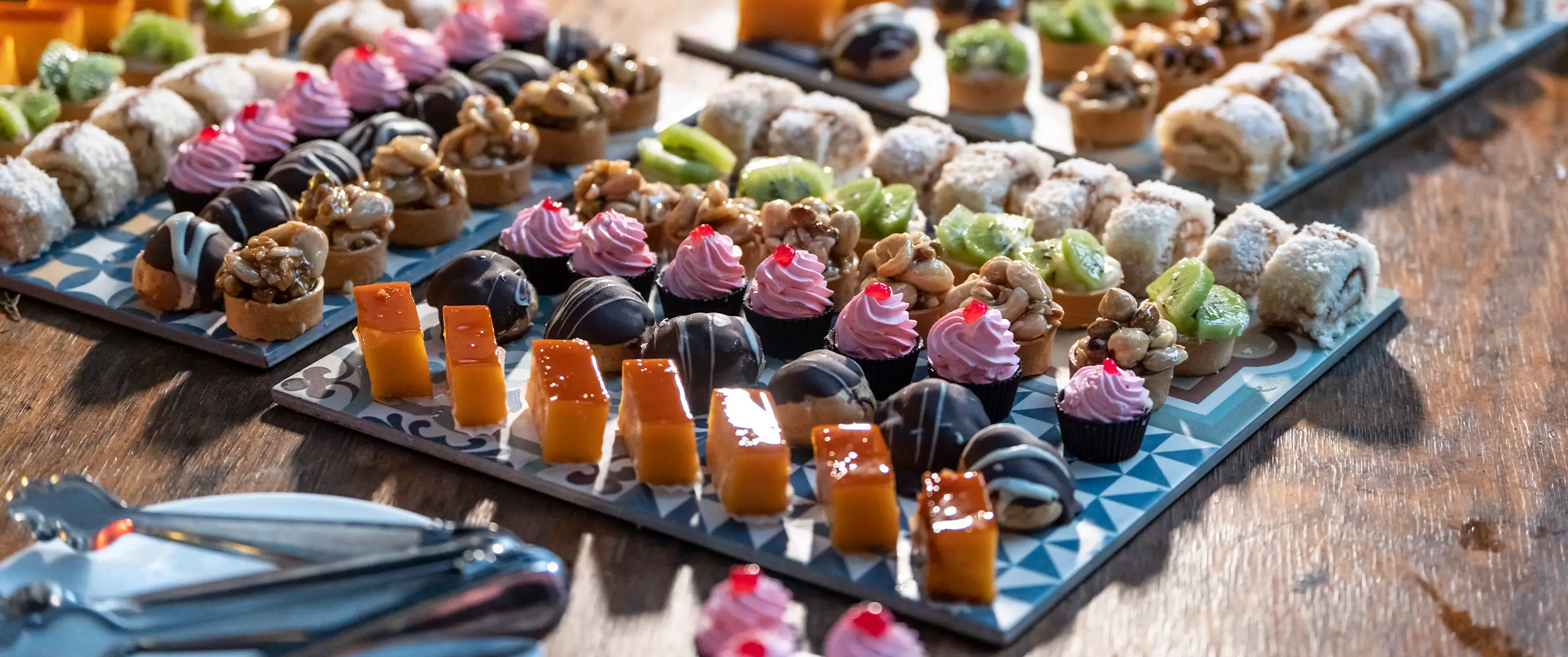 A variety of assorted mini desserts, including pastries, cakes, and tarts, arranged on decorative trays on a wooden table.