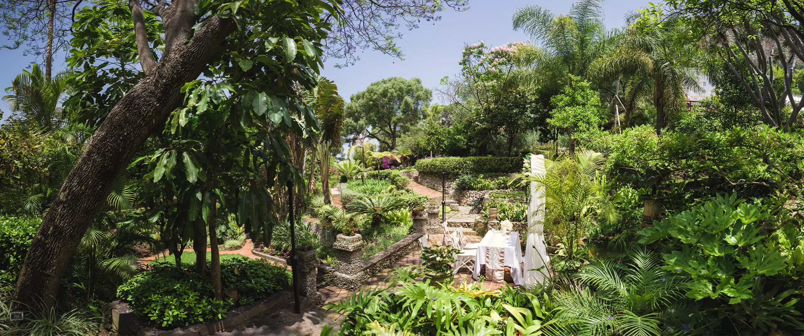 A lush garden with stone pathways, dense green foliage, trees, and a white outdoor table and chairs set among the plants.