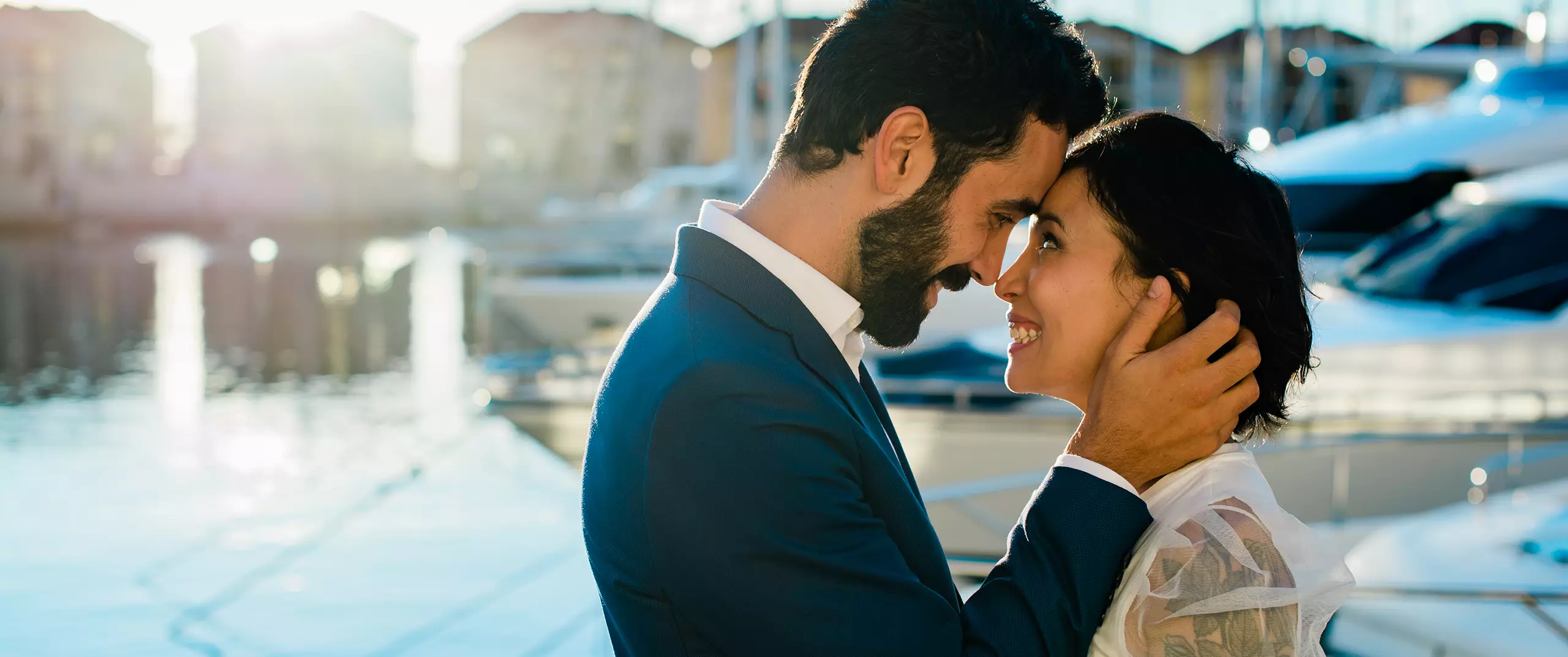 A man and woman in formal attire stand close together by a marina, smiling at each other with their foreheads touching, at sunset.