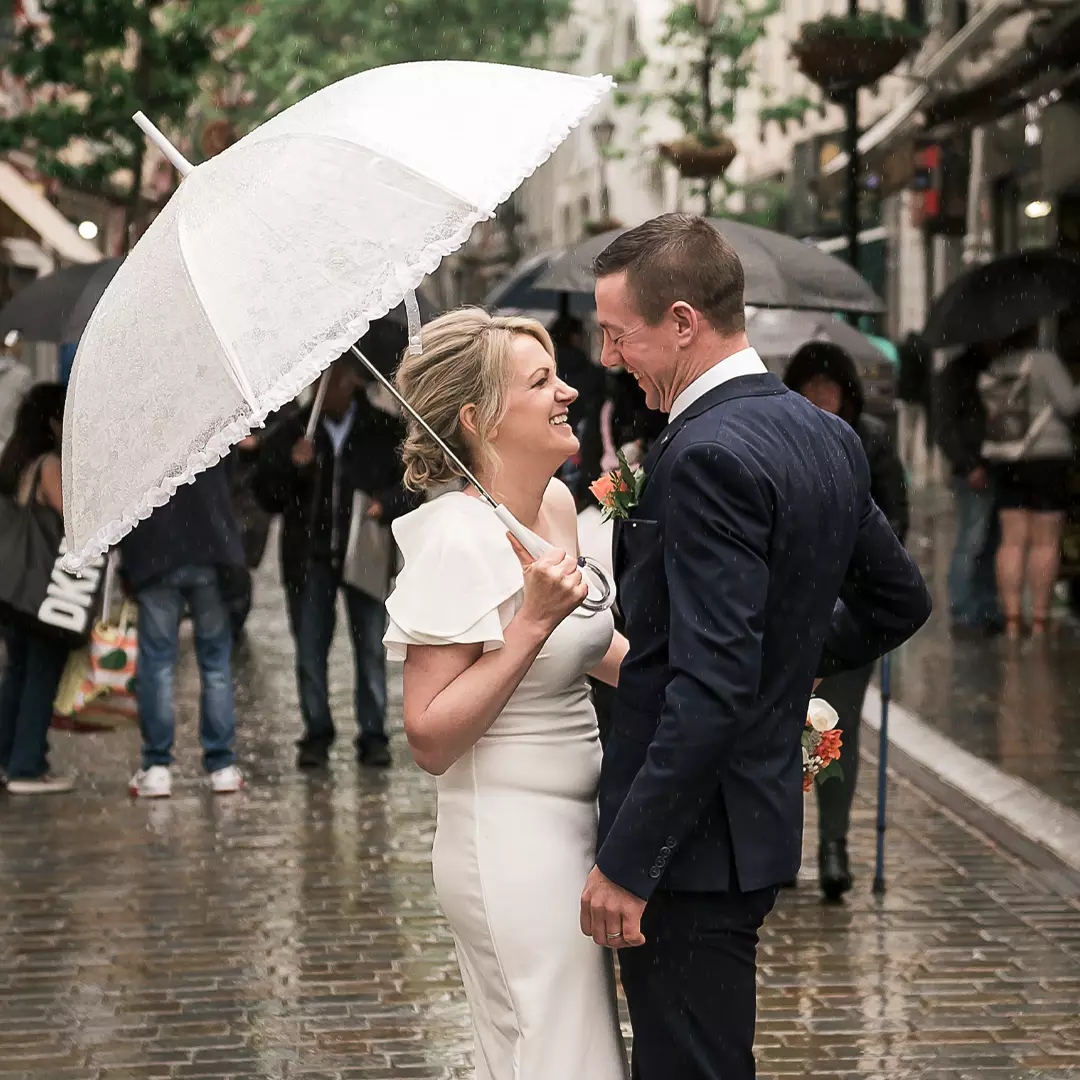 A bride and groom stand under a white umbrella on a rainy street, smiling at each other, surrounded by people with umbrellas in the background.