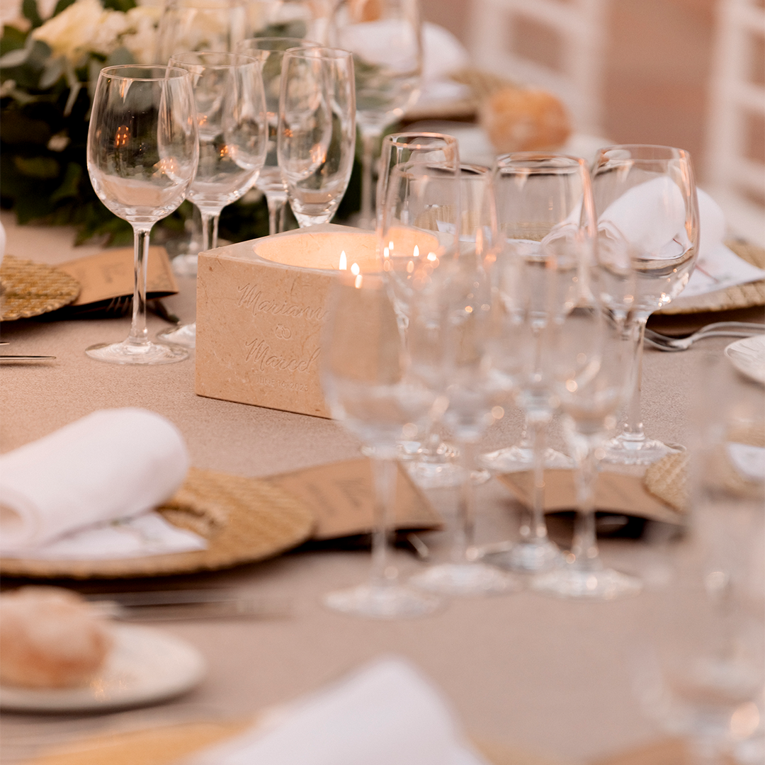 Close-up of an elegant table setting with multiple wine glasses, white napkins on woven placemats, a lit candle, and a floral centerpiece in the background.