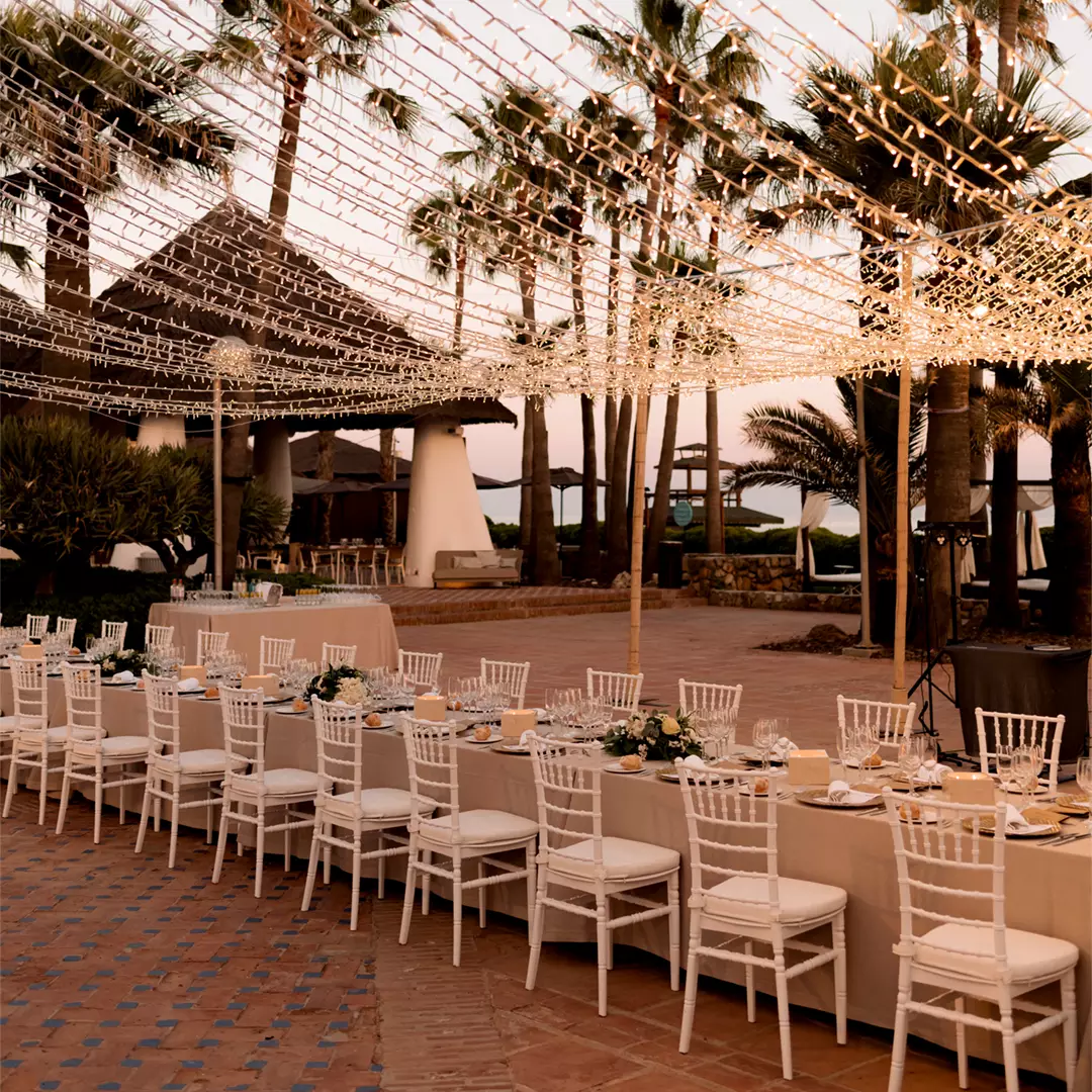 Long banquet tables with white chairs are set for an outdoor event under string lights, surrounded by palm trees and a thatched-roof structure at sunset.