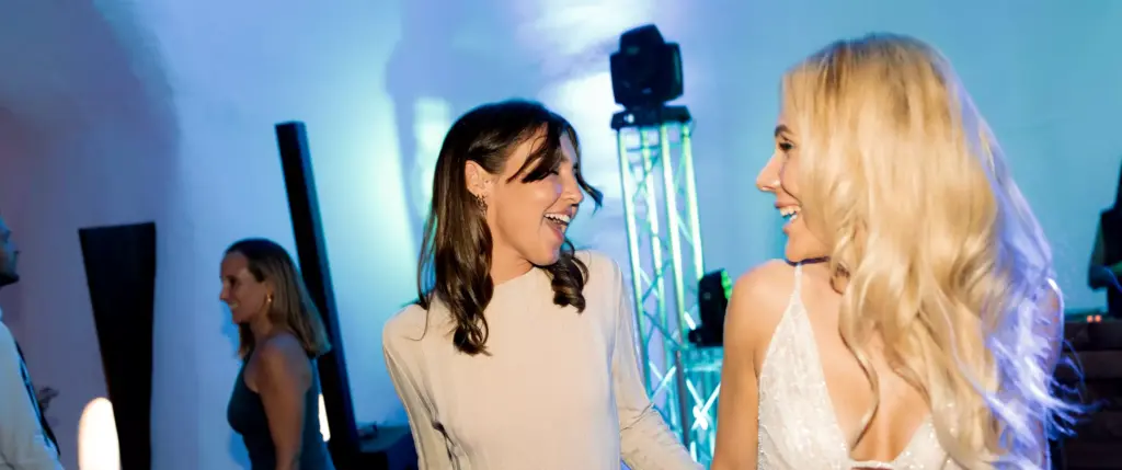 Two women smiling and dancing at an indoor event, with blue lighting and other people in the background.