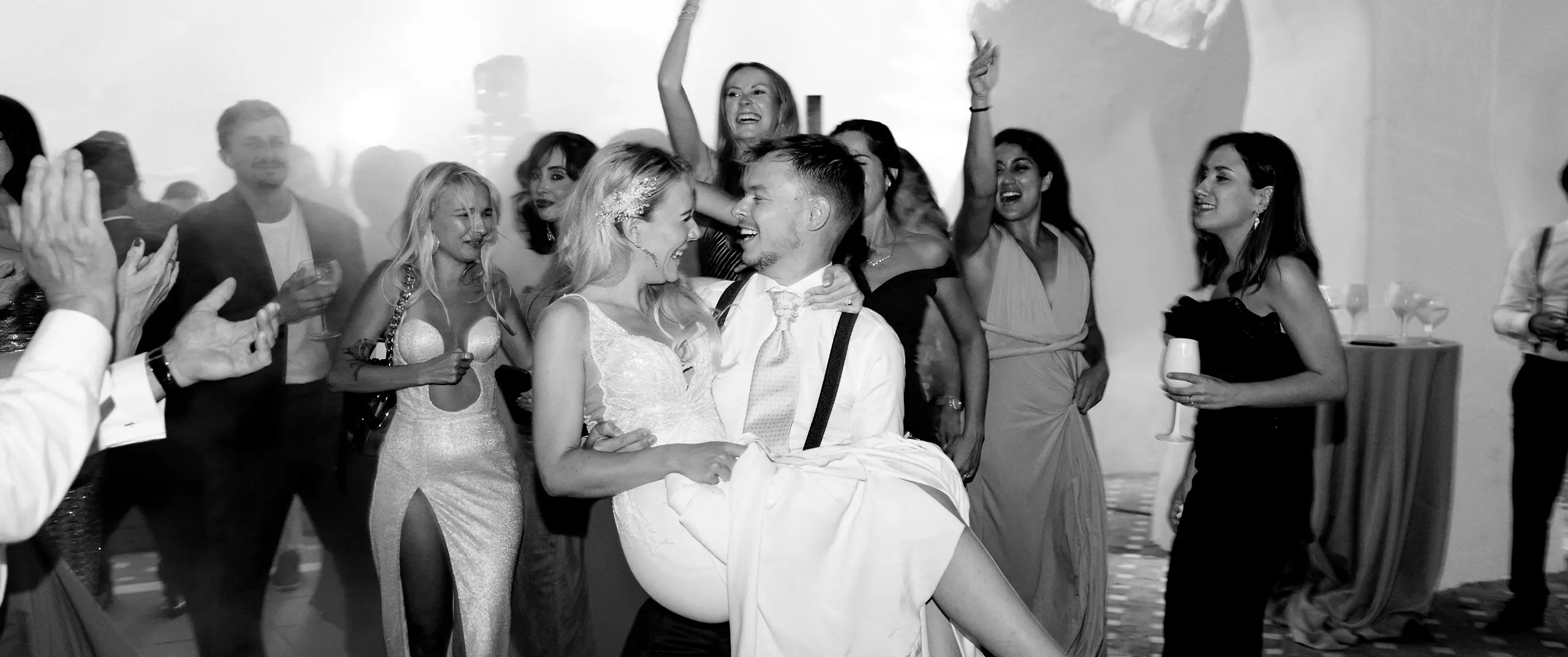 A groom holds his bride while surrounded by friends celebrating and cheering at a wedding reception.