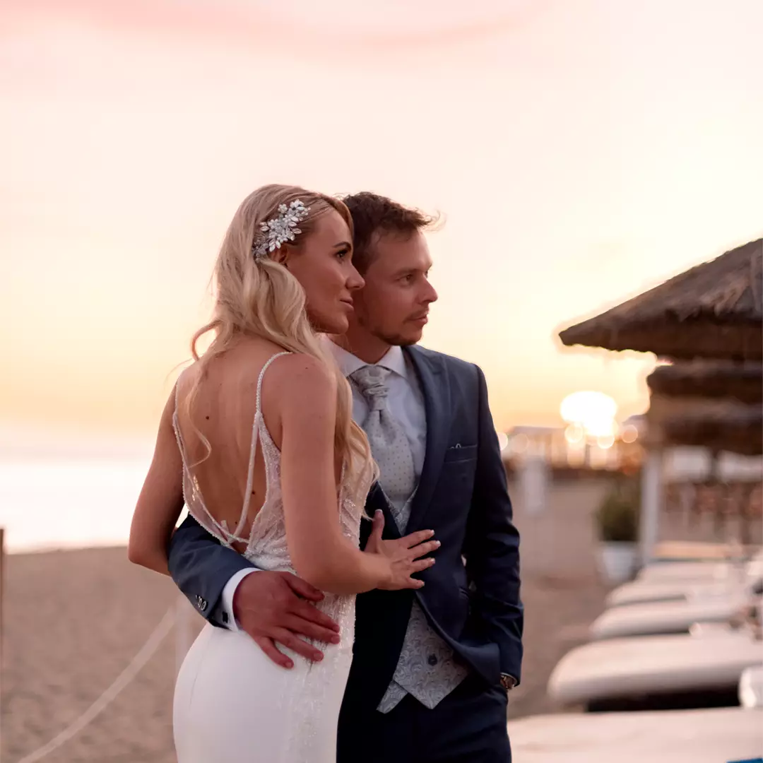 A bride and groom stand close together on a beach at sunset, dressed in formal wedding attire, looking into the distance.