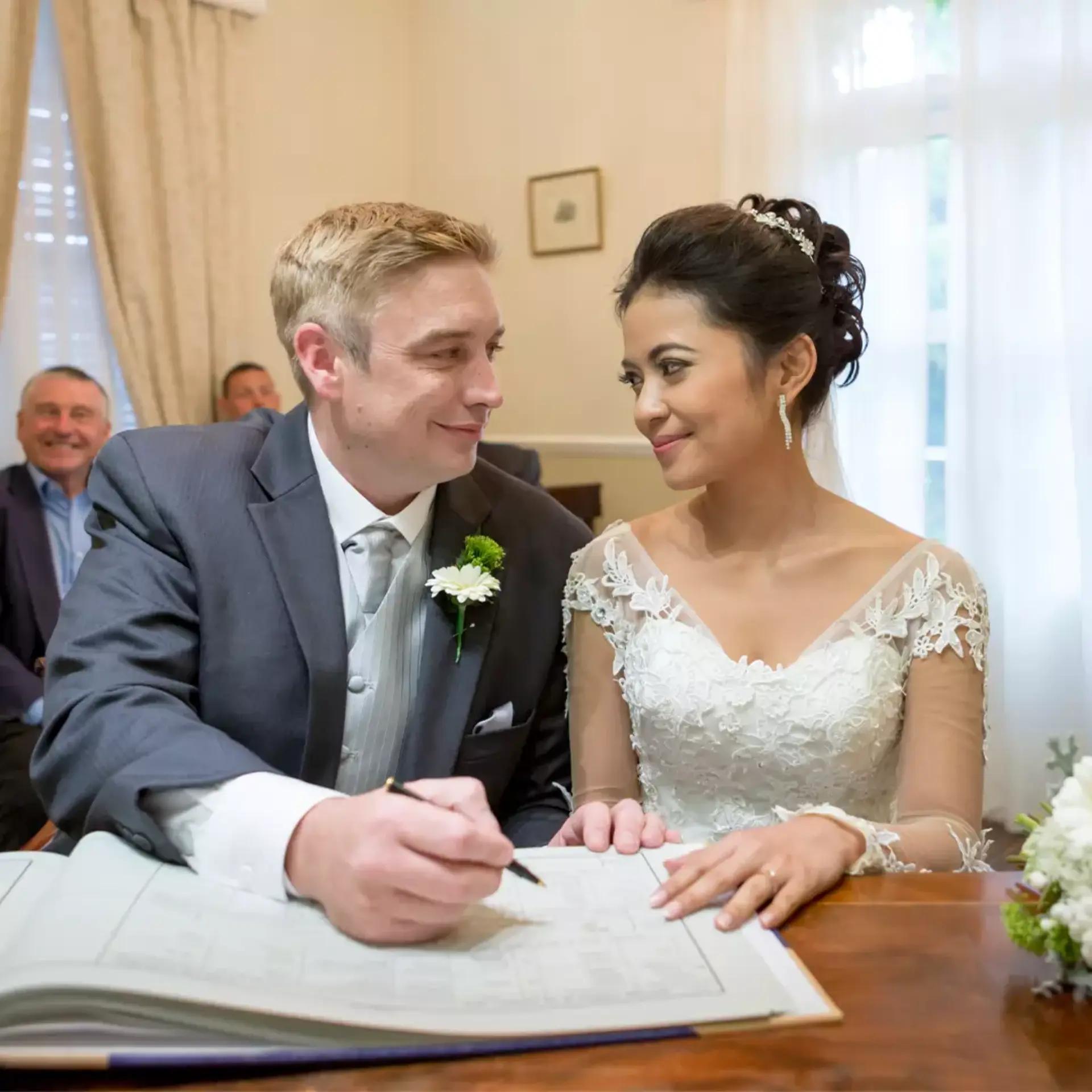 A bride and groom sit together at a table, smiling at each other as the groom signs a document, likely at a wedding ceremony, with guests visible in the background.