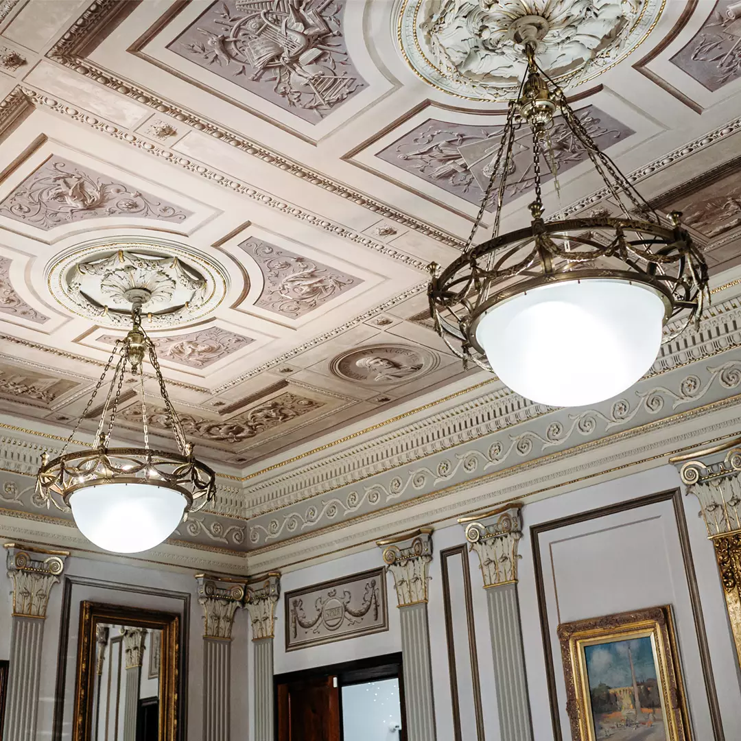 Ornate ceiling with decorative molding and two large, round chandeliers; classic framed paintings and mirrors line the walls below.