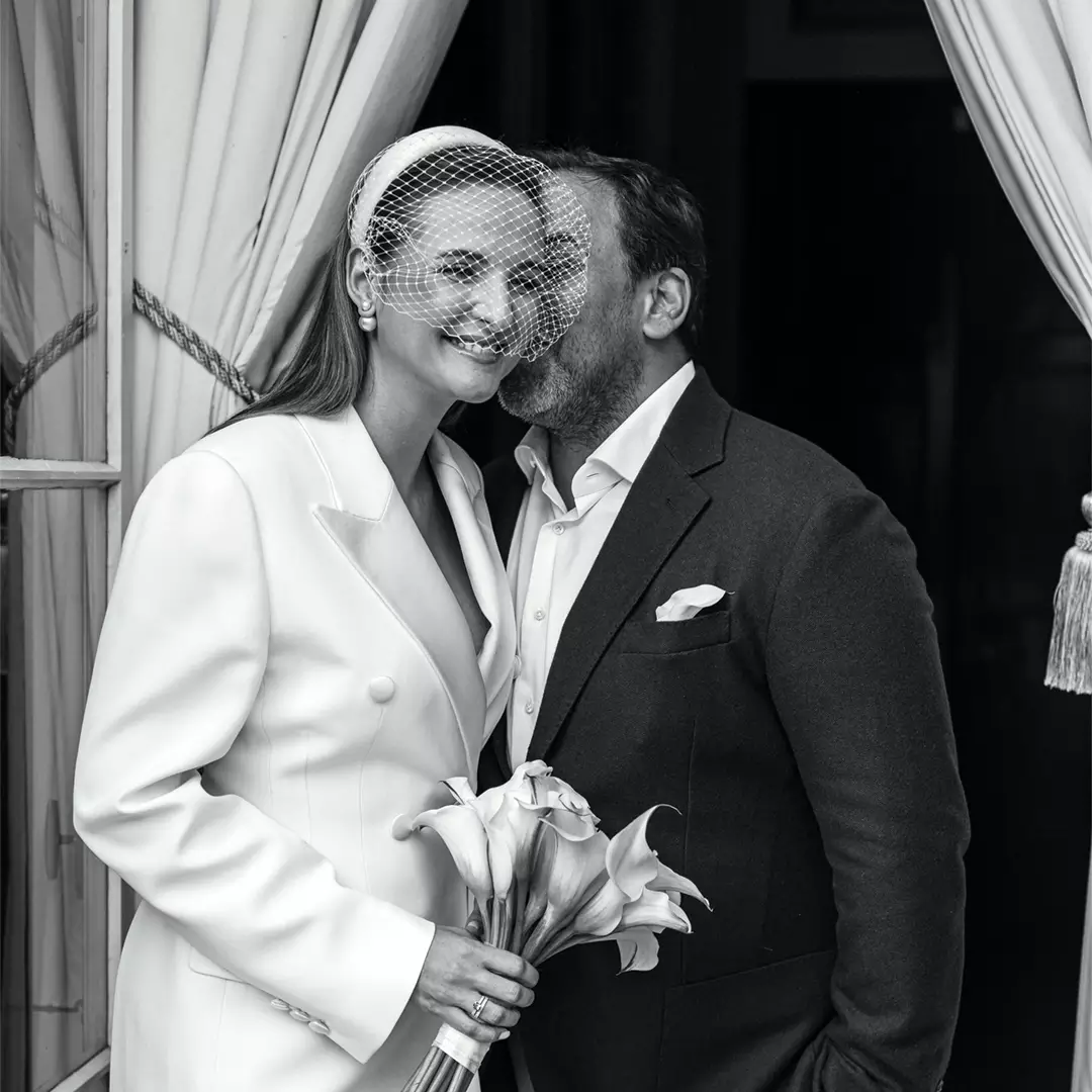 A woman in a white suit and veil holds calla lilies while smiling next to a man in a suit who is whispering in her ear; they stand by an open curtain.