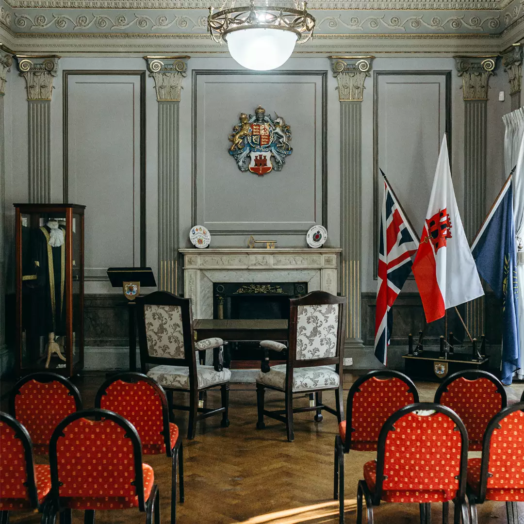 Ornate room with red-upholstered chairs facing two armchairs, a fireplace, three flags, a coat of arms, and traditional garments displayed in a glass case.