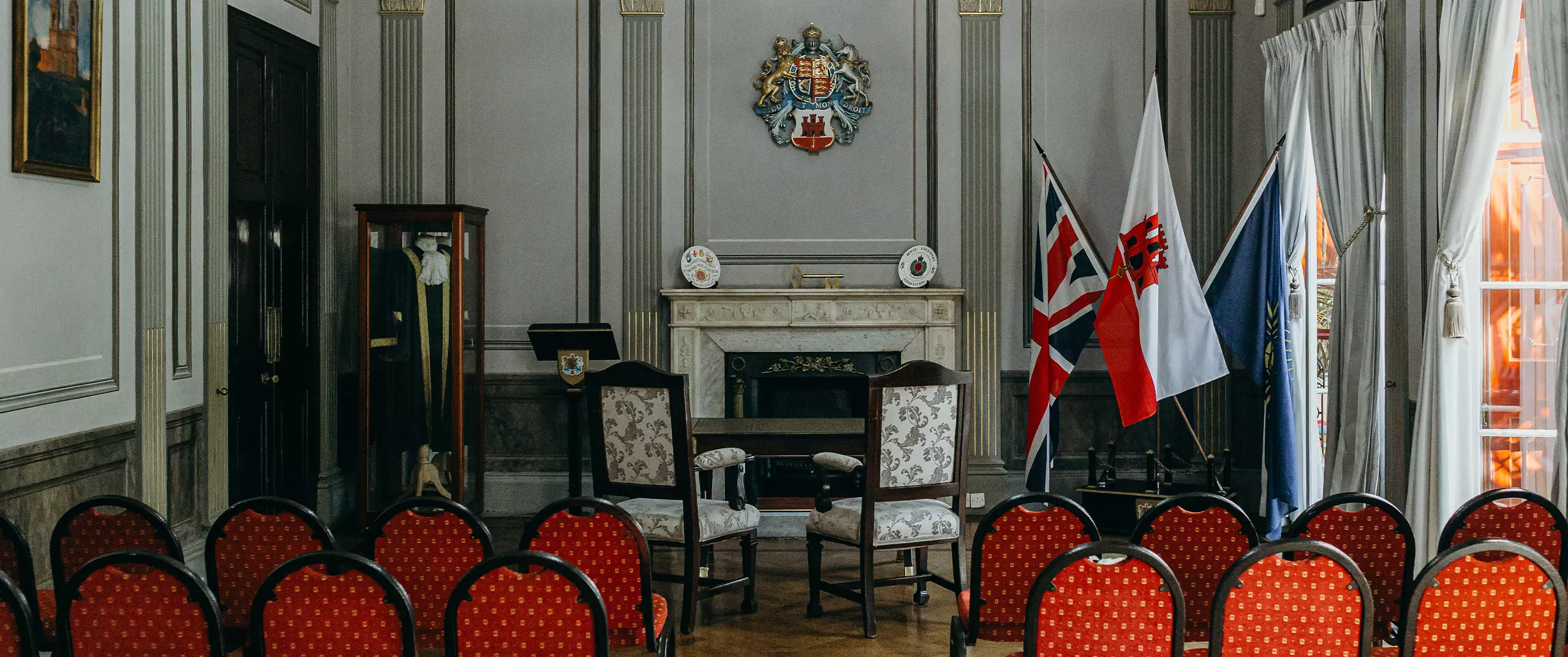 A formal room with red chairs arranged facing a fireplace, two armchairs, national flags, and a coat of arms above the mantel.
