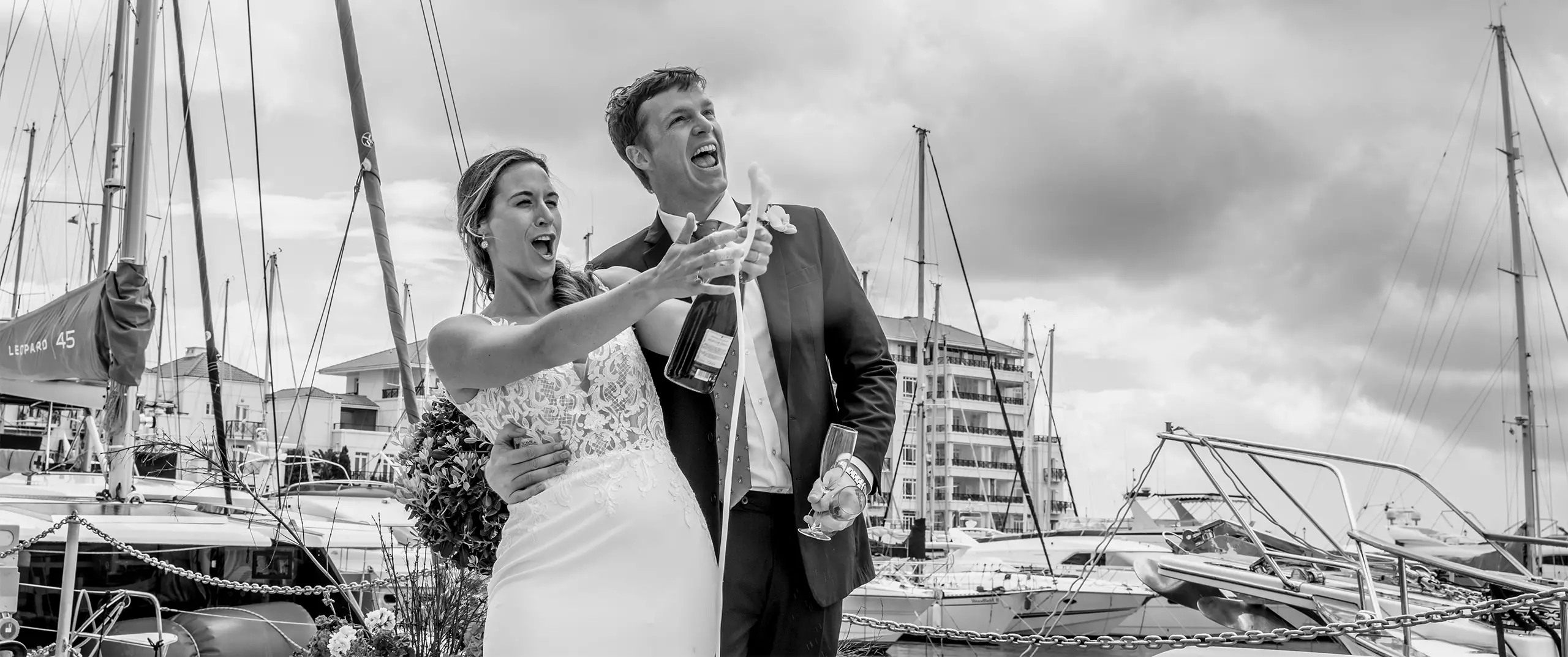 A bride and groom, dressed in wedding attire, celebrate and open a bottle of champagne at a marina with boats and cloudy skies in the background.