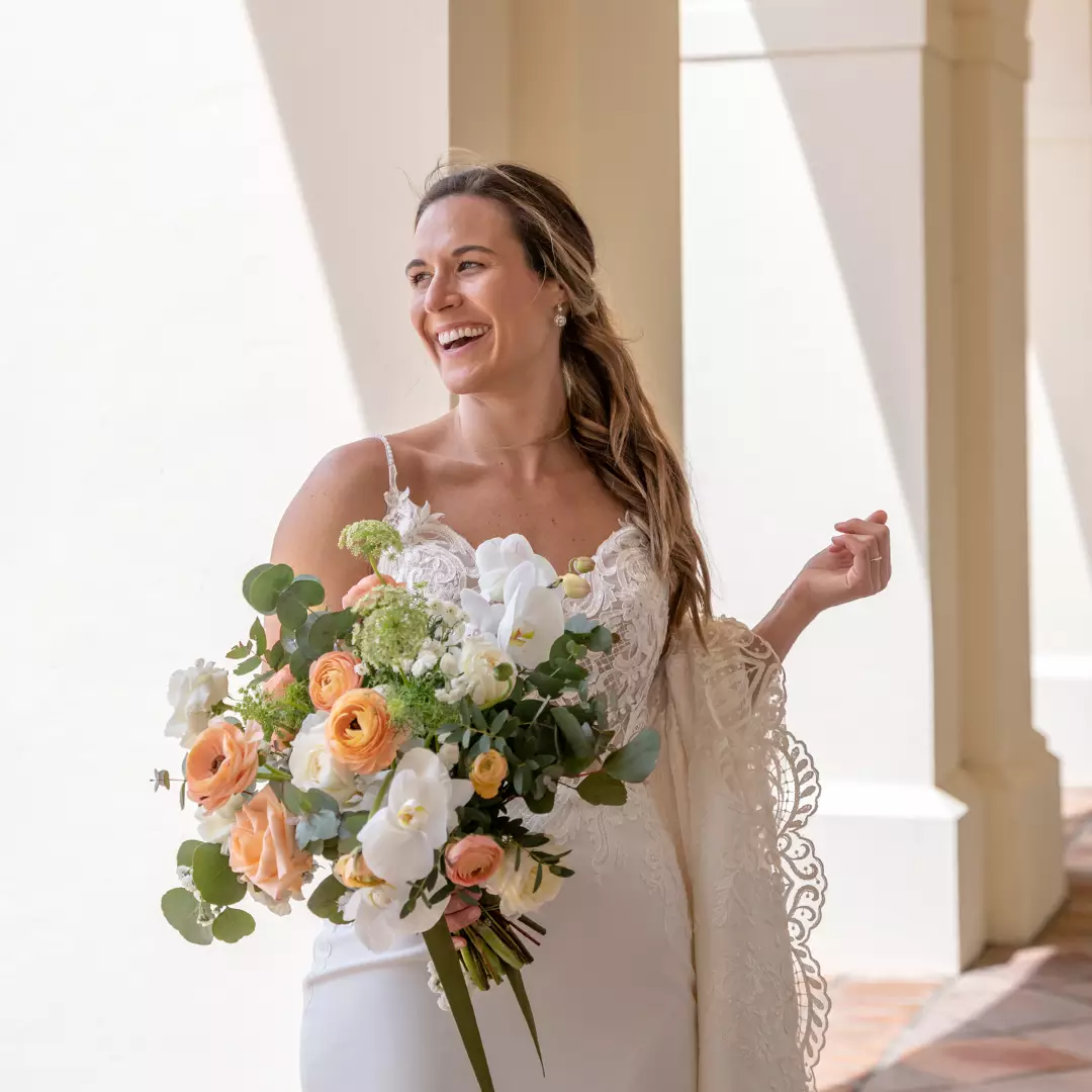 A bride in a white lace wedding dress holds a bouquet of flowers and smiles while standing in a sunlit corridor.
