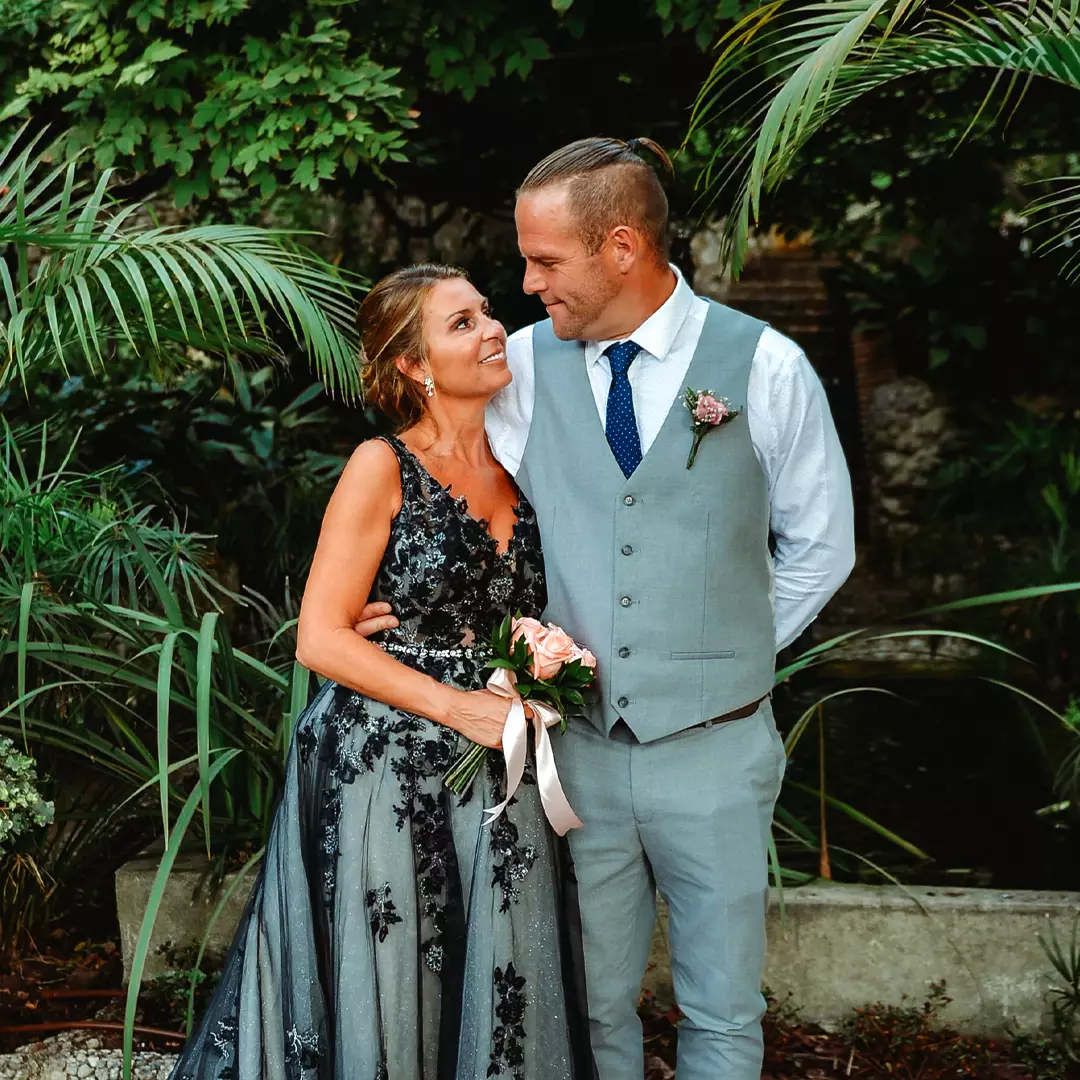 A woman in a dark floral dress and a man in a light gray vest and tie stand close together, holding a bouquet, and smiling at each other in an outdoor garden setting.
