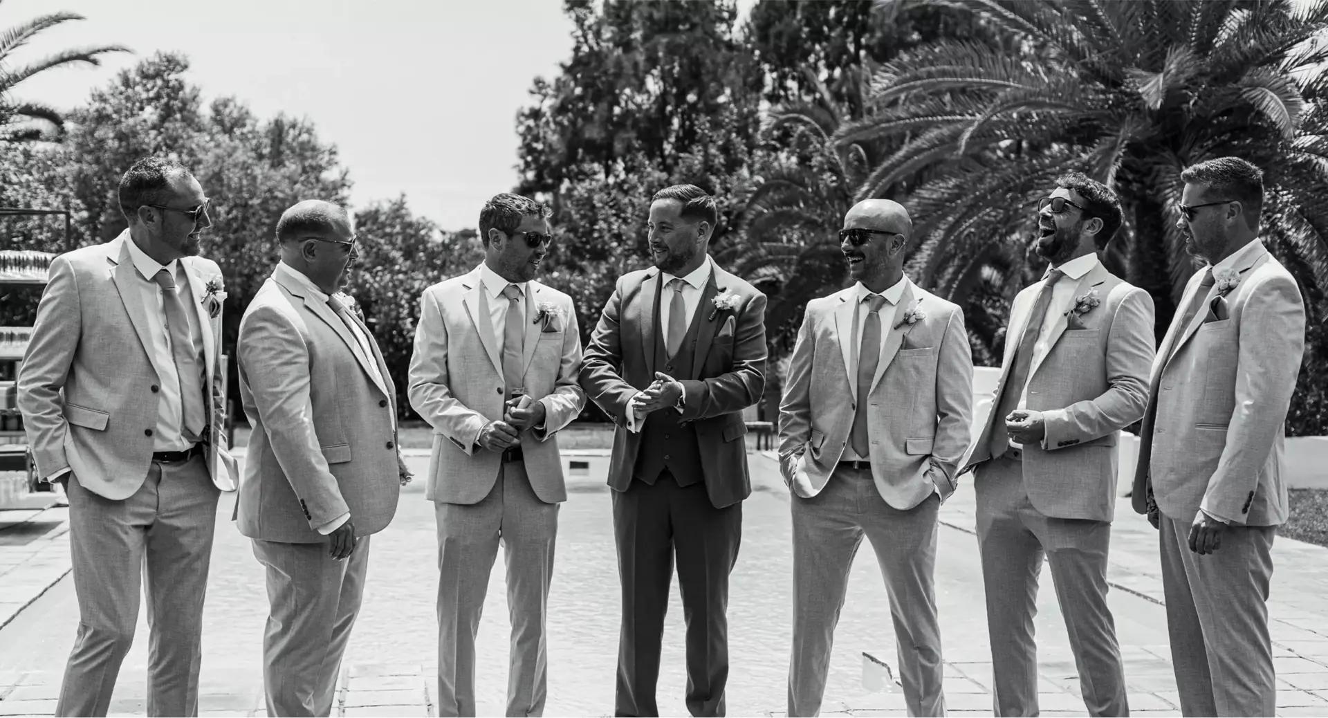 Six men in suits stand together outdoors near a pool, talking. The photo is black and white, with trees and palm fronds in the background.