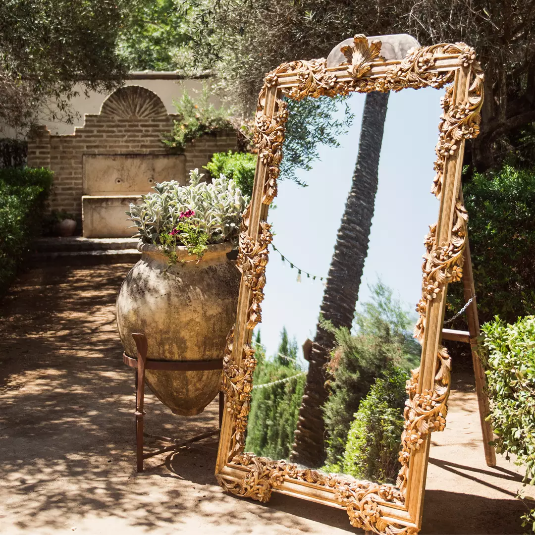 An ornate gold-framed mirror stands outdoors on a path, reflecting a tall palm tree and greenery; a large planter with flowers is beside it.