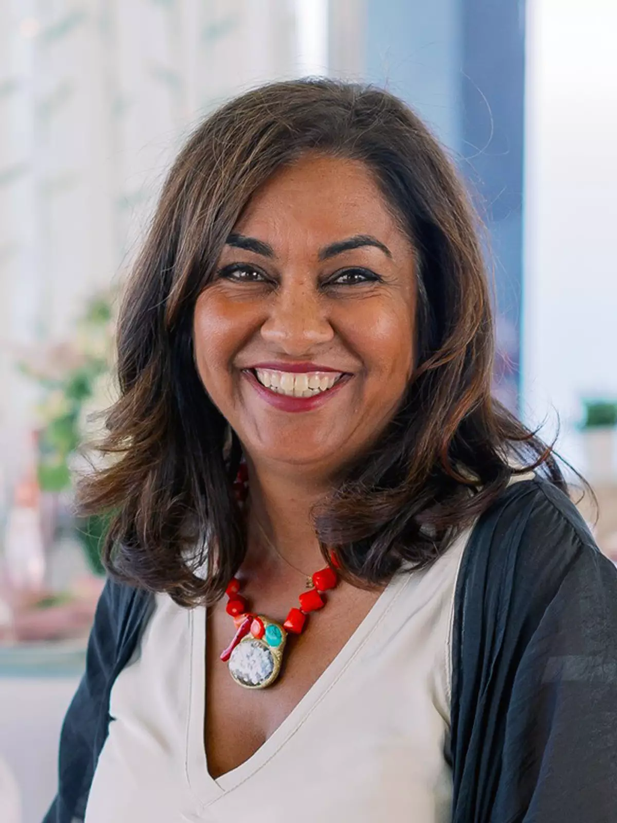 A woman with wavy brown hair smiles at the camera, wearing a white top, a black shawl, and a bold red necklace.