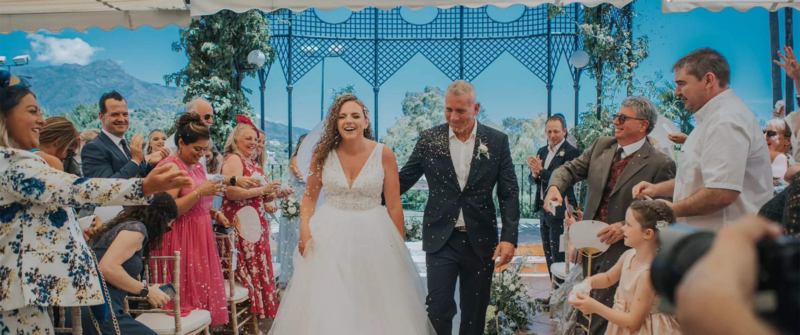A bride and groom walk down the aisle outdoors, smiling as guests celebrate and throw confetti; mountains and greenery are visible in the background.