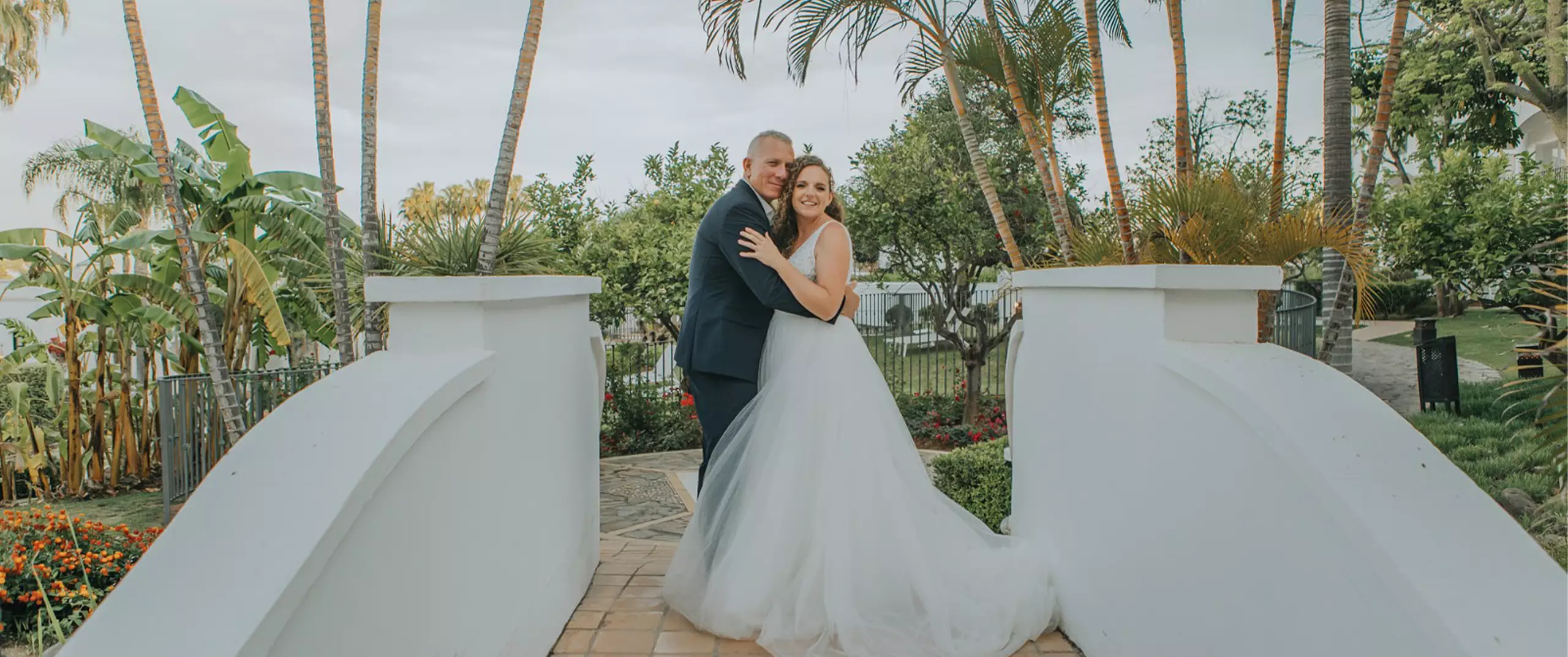 A bride and groom in formal attire embrace on a white pedestrian bridge, surrounded by palm trees and lush greenery.