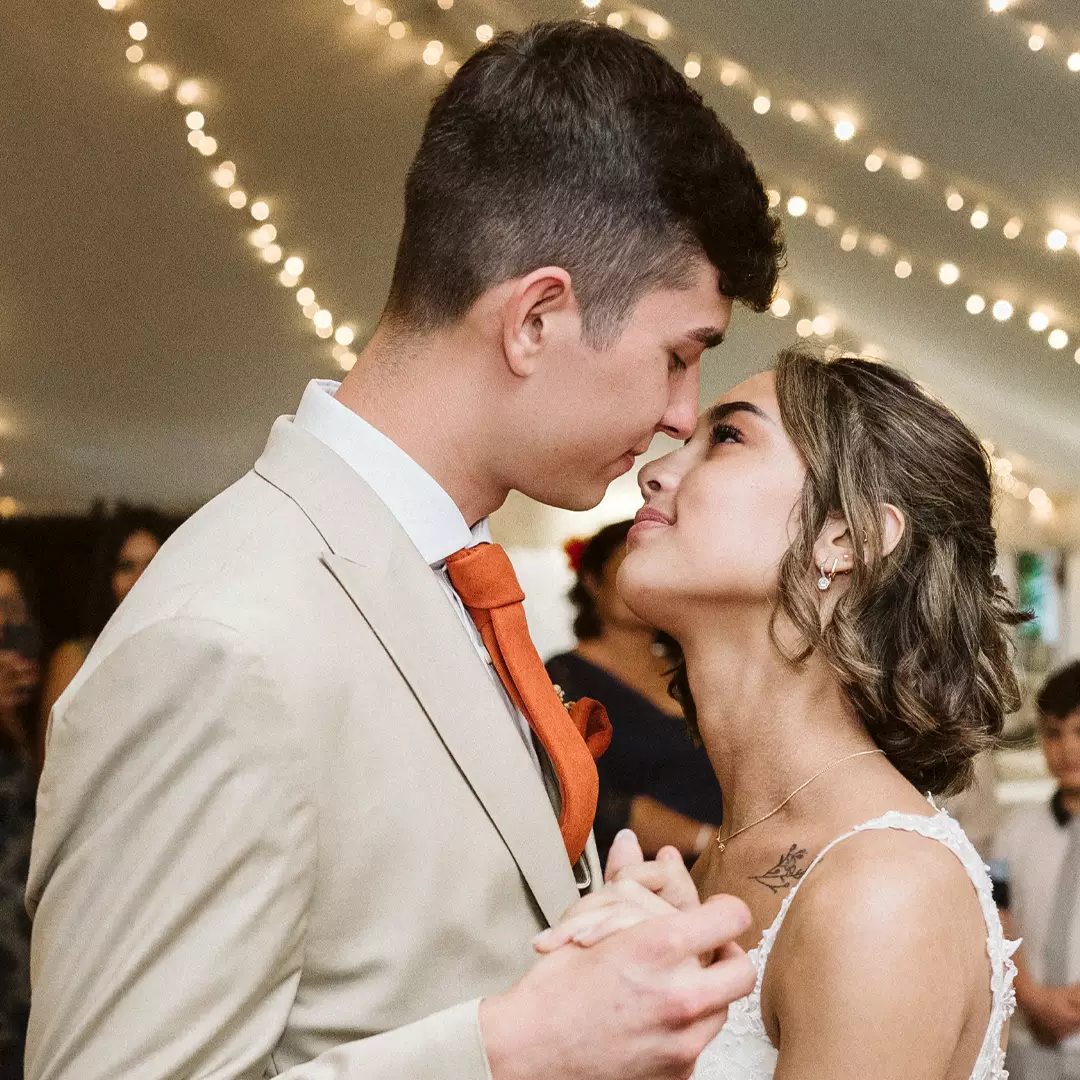 A bride and groom dance closely under string lights, gazing into each other's eyes while holding hands at their wedding reception.