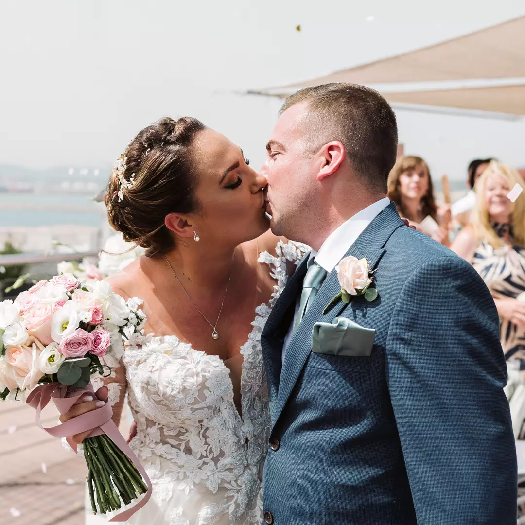 A bride and groom kiss outdoors on their wedding day. The bride holds a bouquet of flowers, and guests watch in the background.
