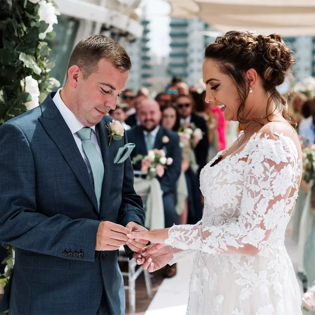 A groom places a ring on the bride's finger during a wedding ceremony, with guests watching in the background. Both are dressed in formal attire.