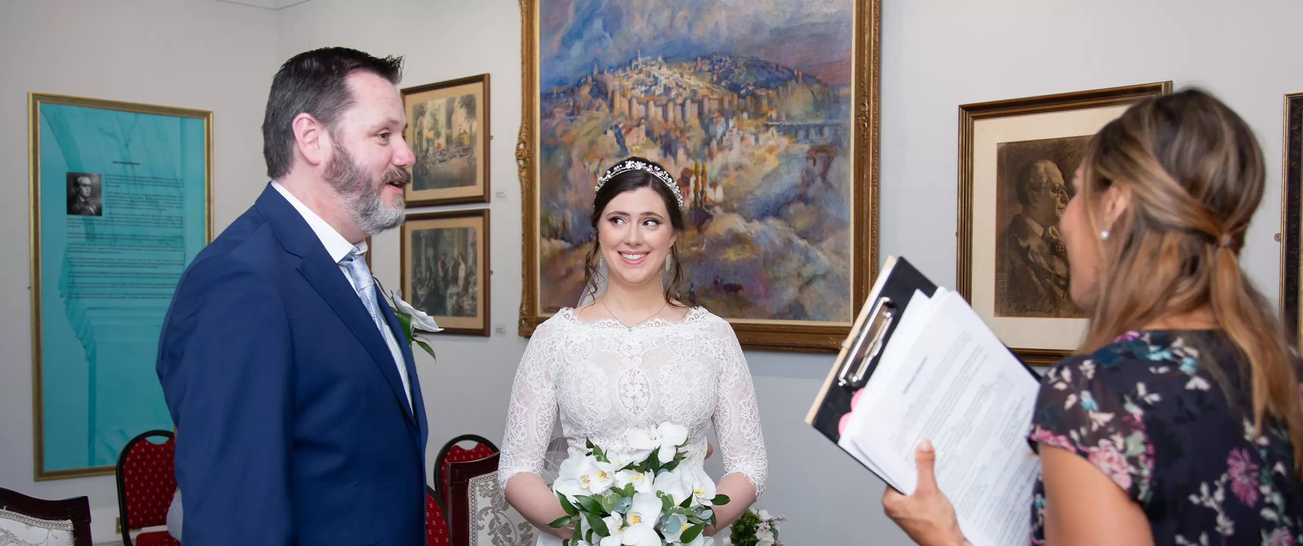 A bride and groom stand together indoors, facing a woman holding a clipboard, possibly during a wedding ceremony. Paintings and framed art are visible on the walls behind them.