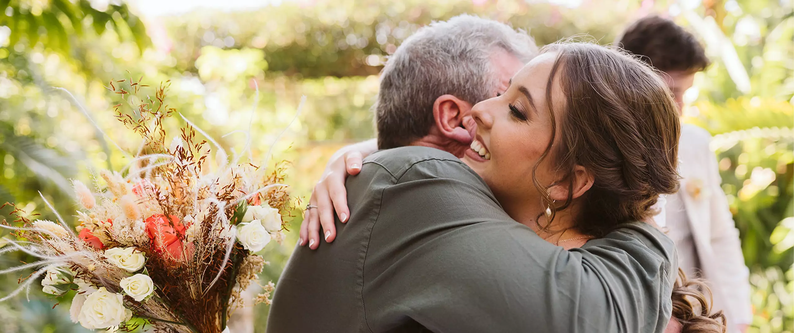 A woman smiling and hugging a man outdoors, with a bouquet of flowers visible on the left side of the image.
