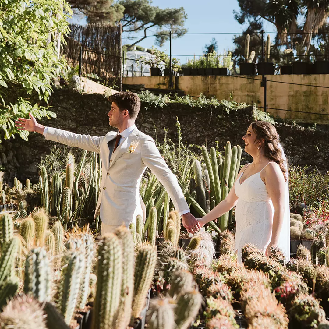 A bride and groom in wedding attire hold hands and walk through a garden with cacti and succulents, with the groom extending his free arm outward.