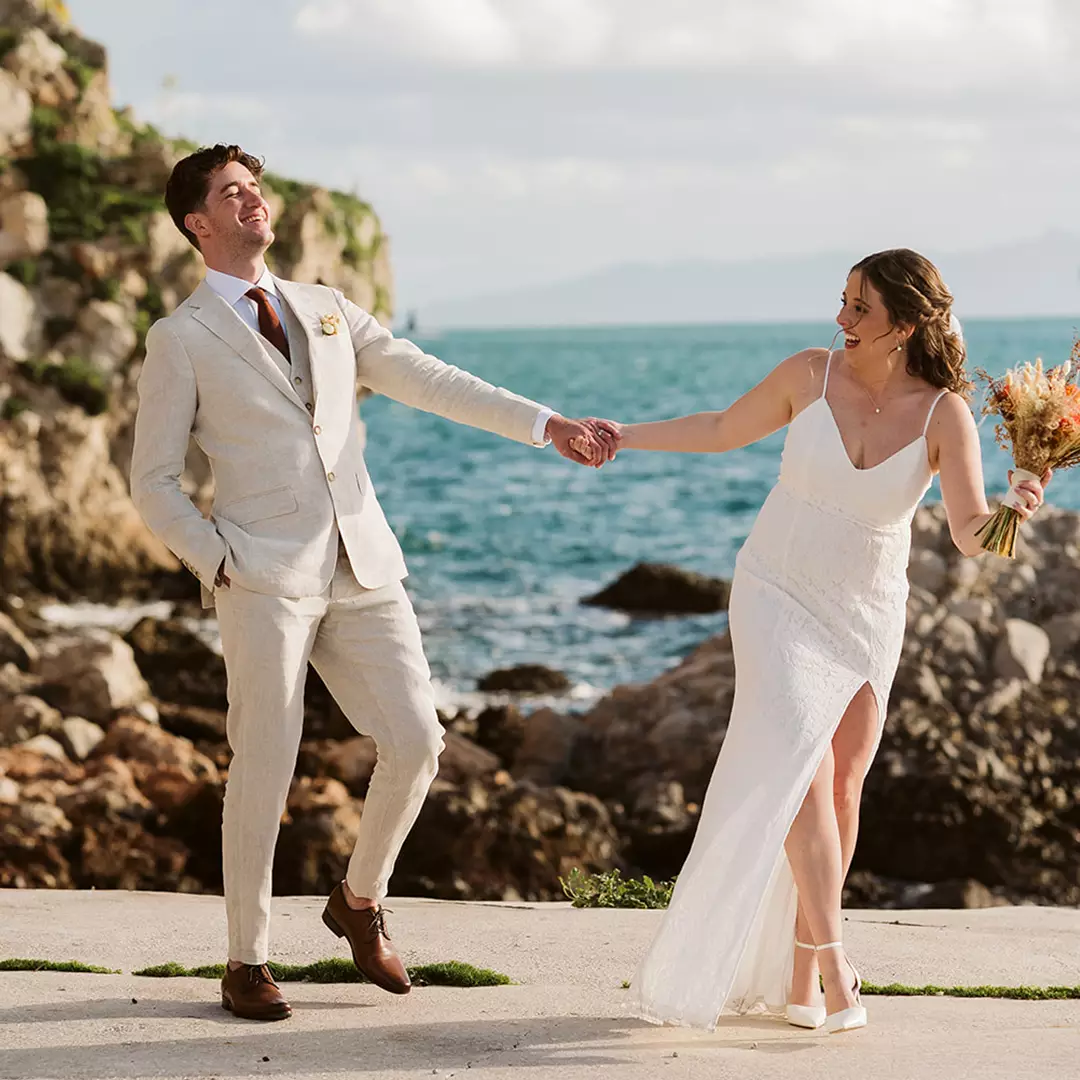 A bride and groom hold hands and smile at each other while standing near the ocean on a rocky shoreline.