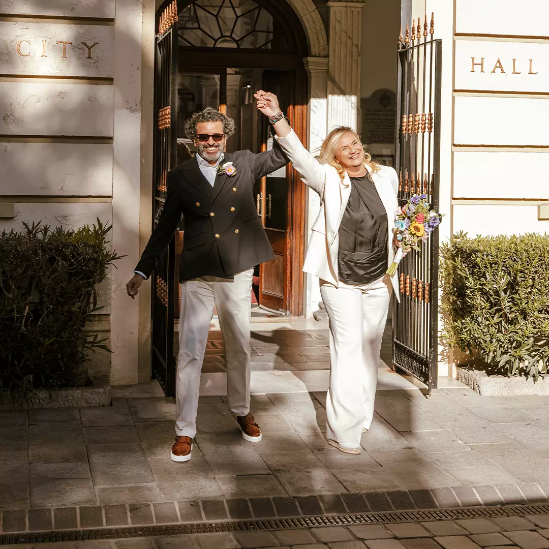 A smiling couple holding hands and celebrating as they exit a city hall building, with one holding a bouquet of flowers.