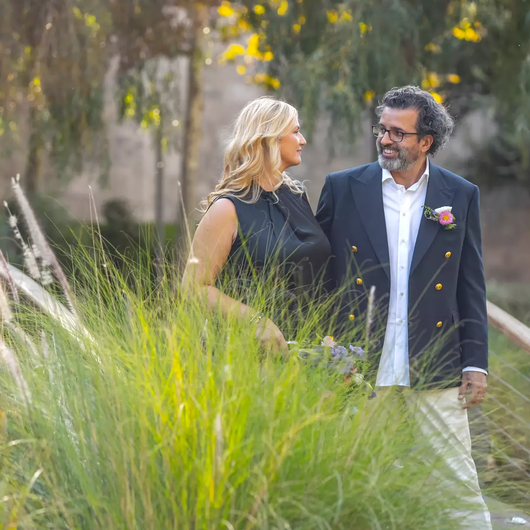 A man and woman stand together outdoors, smiling at each other, surrounded by tall green grass and trees in the background.