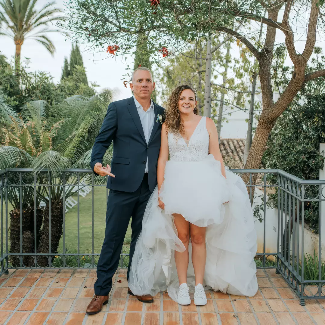 A bride in a white dress and sneakers stands next to a man in a navy suit on a tiled patio, with greenery and trees in the background.