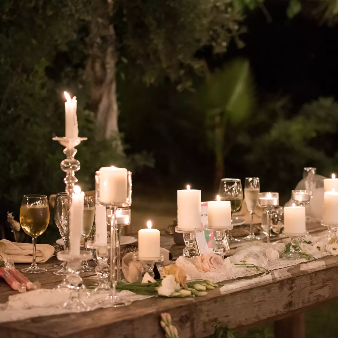 A rustic wooden table outdoors is decorated with lit candles, wine glasses, flowers, and lace, creating an elegant, ambient setting at night.