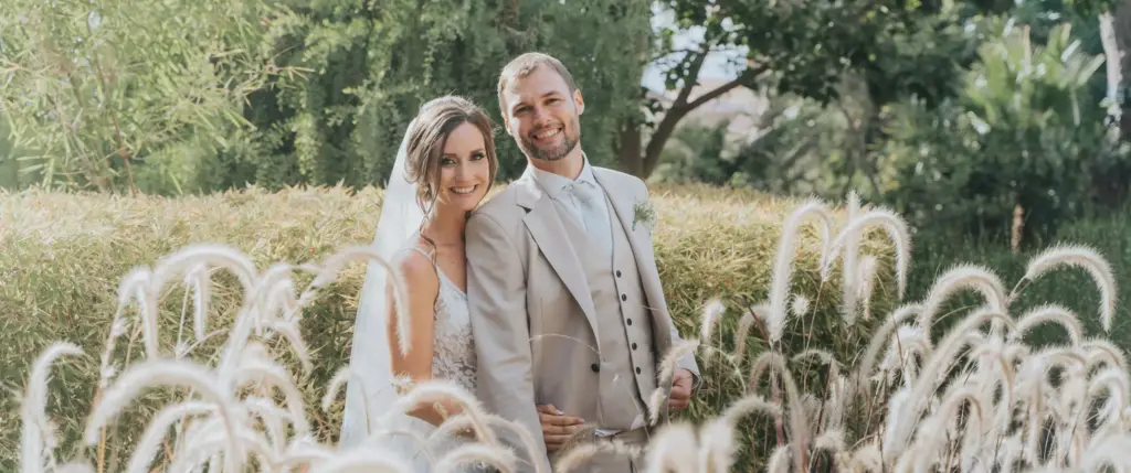 Bride and groom smiling and posing together outdoors in front of greenery and ornamental grass on their wedding day.