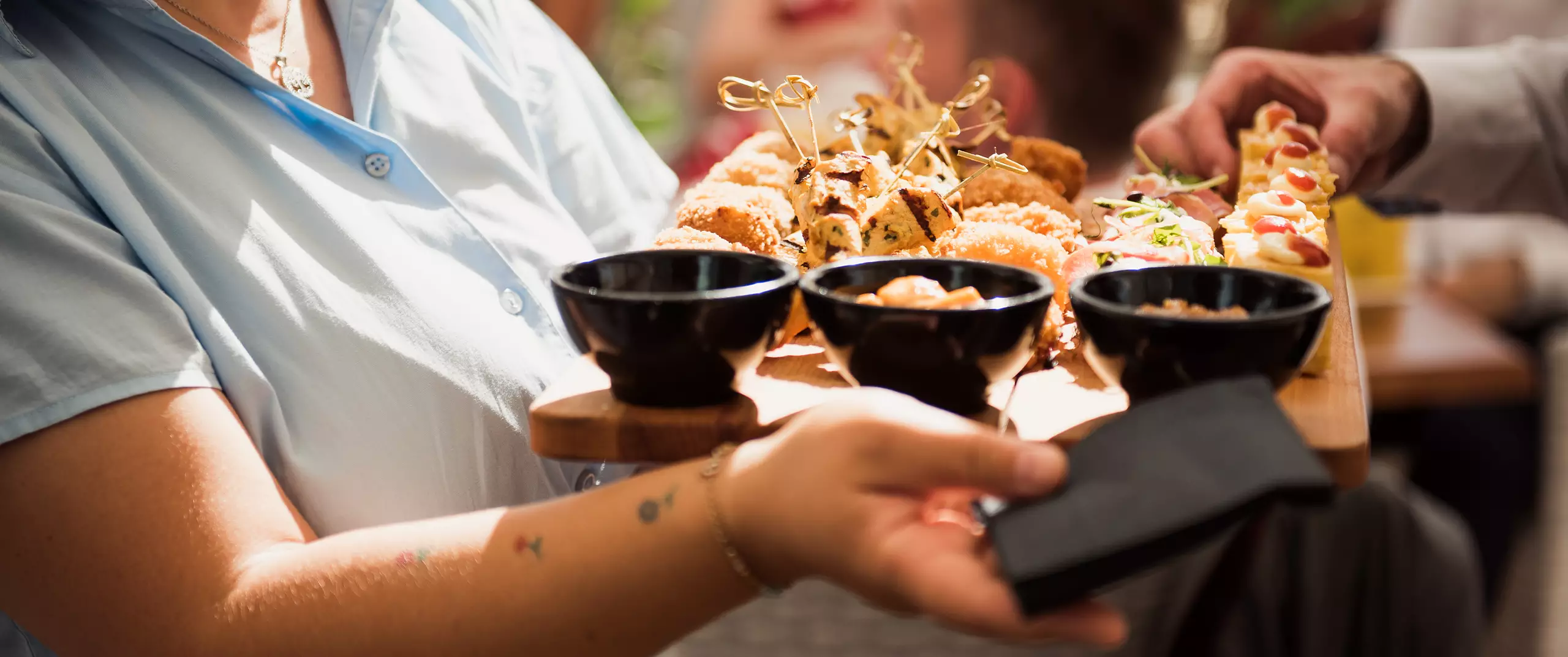 A person holds a wooden tray with three black bowls and assorted appetizers while another hand reaches for food.