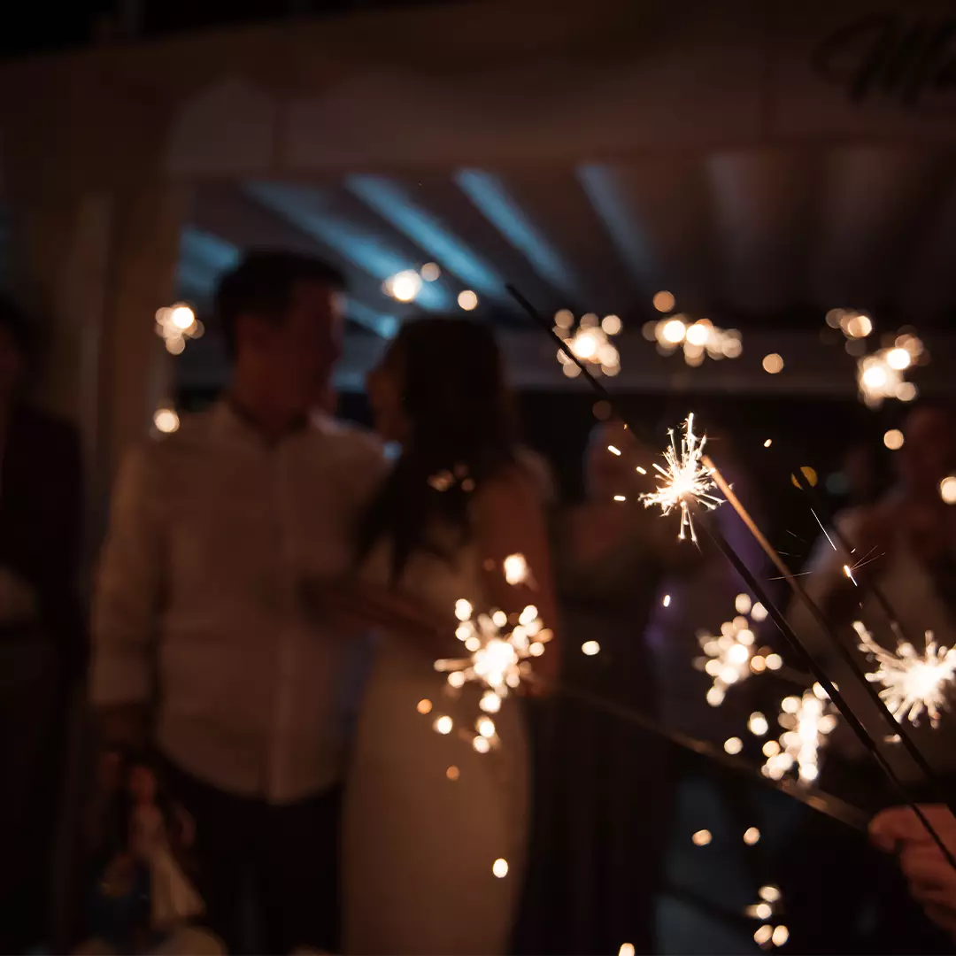 A group of people hold sparklers at night, with a couple standing close together in the background.