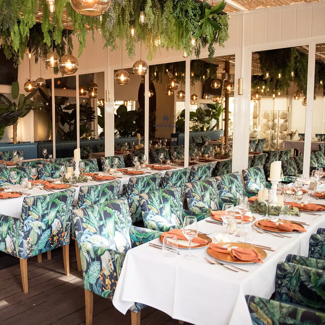 A restaurant dining area with floral-patterned chairs, white tablecloths, orange plates, and hanging greenery with pendant lights above.