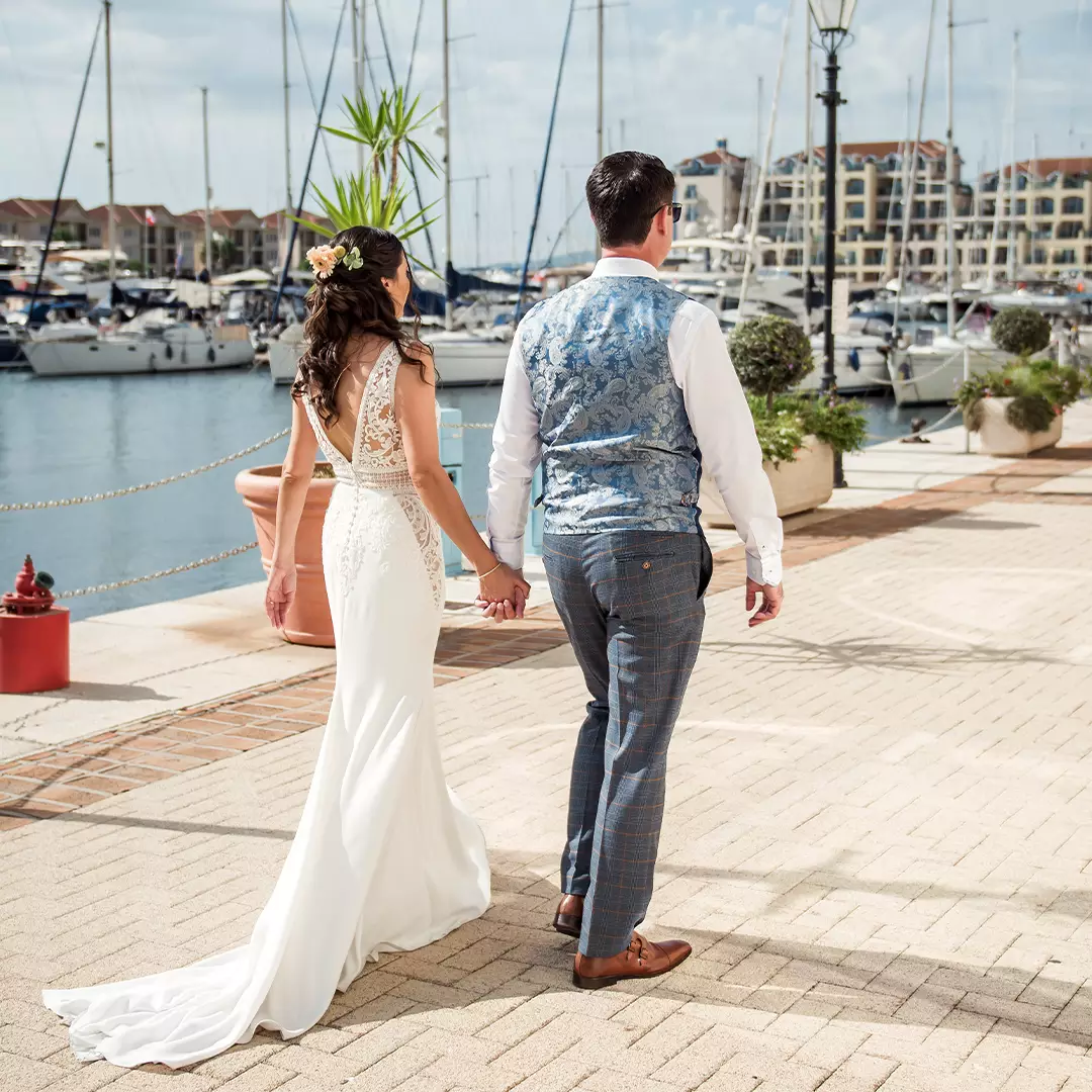 A bride and groom walk hand in hand along a marina, with boats and waterfront buildings in the background.