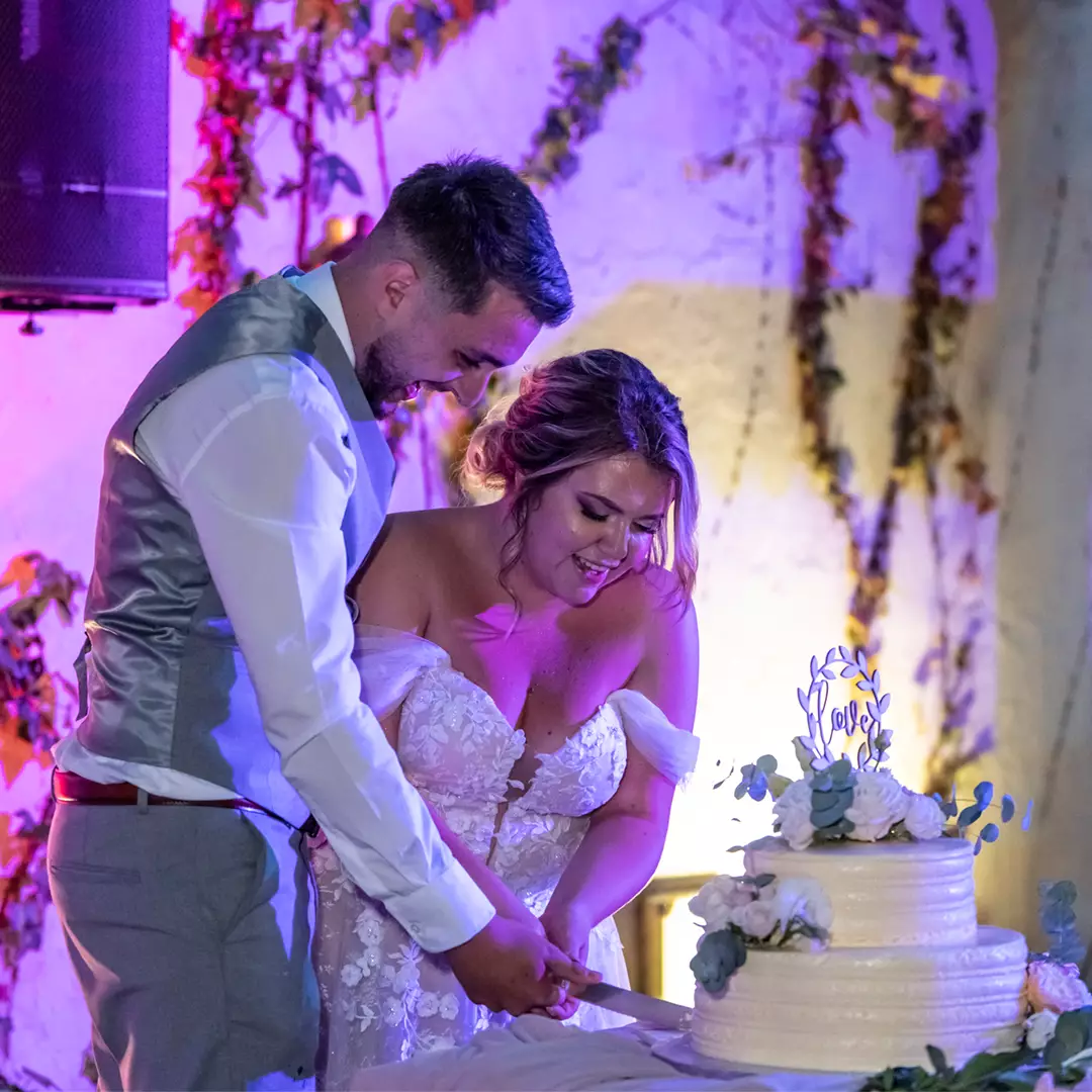 A bride and groom in formal attire cut a white wedding cake together at a decorated indoor venue with greenery on the walls.