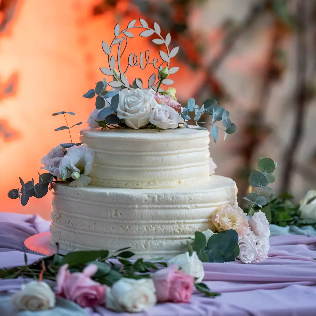 Two-tiered white cake decorated with roses, eucalyptus leaves, and a topper that says 