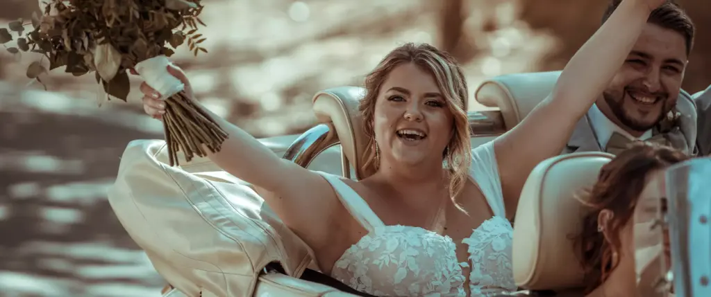 A bride in a white dress smiles and raises her bouquet while sitting in a car next to a man in a suit, with another person partially visible in the foreground.
