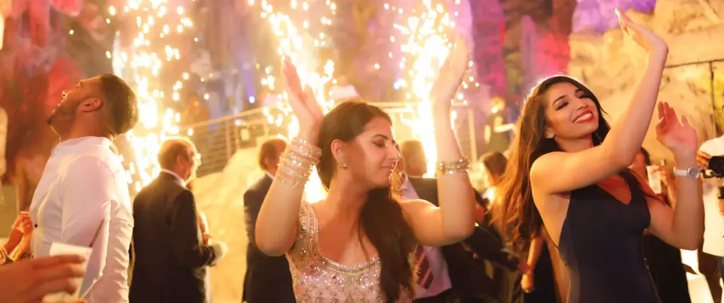 People dancing and celebrating at a party with sparklers in the background, wearing formal attire and smiling.