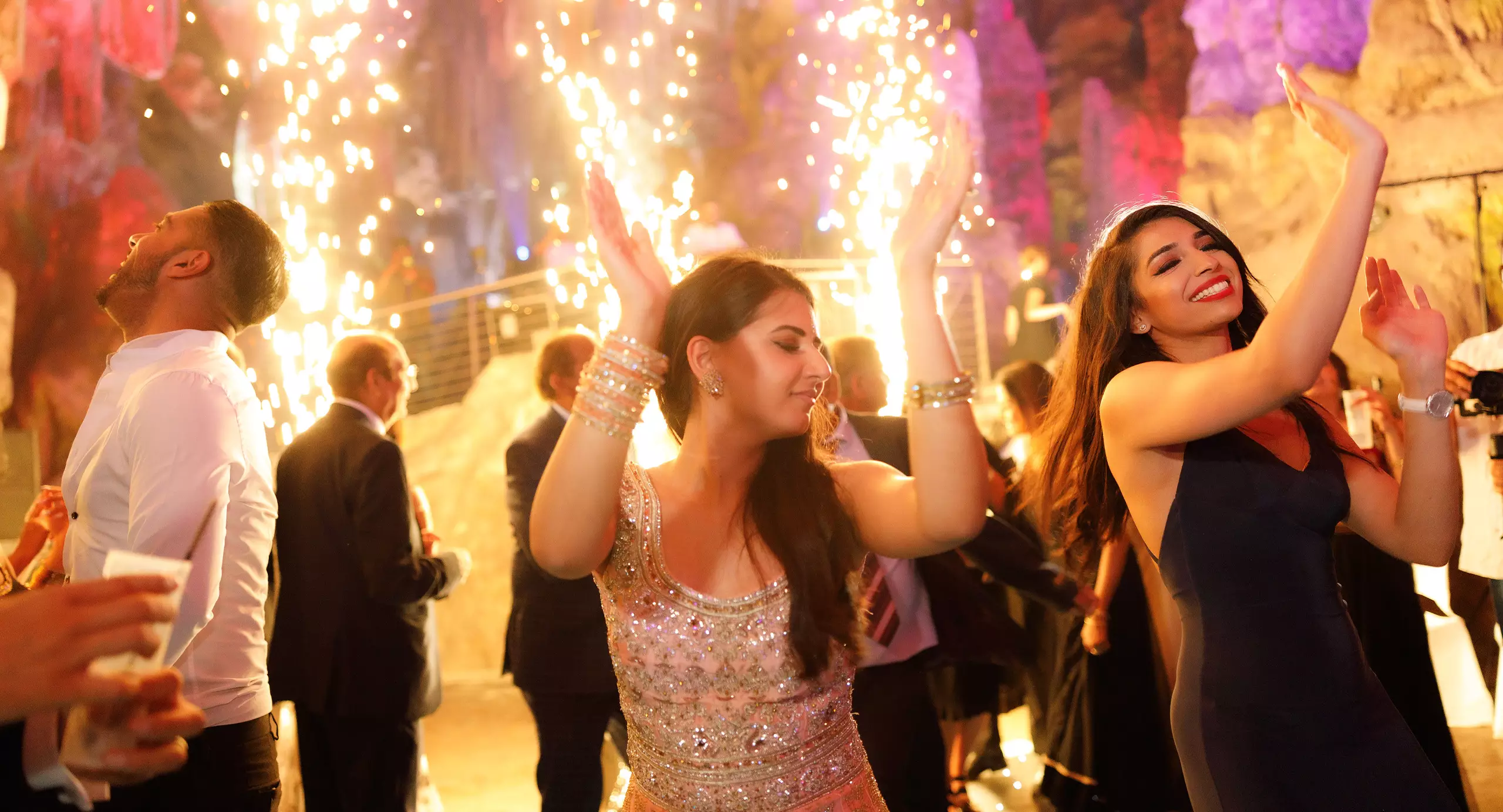 People dressed formally dance and celebrate under indoor fireworks at a lively event with colorful lights in the background.
