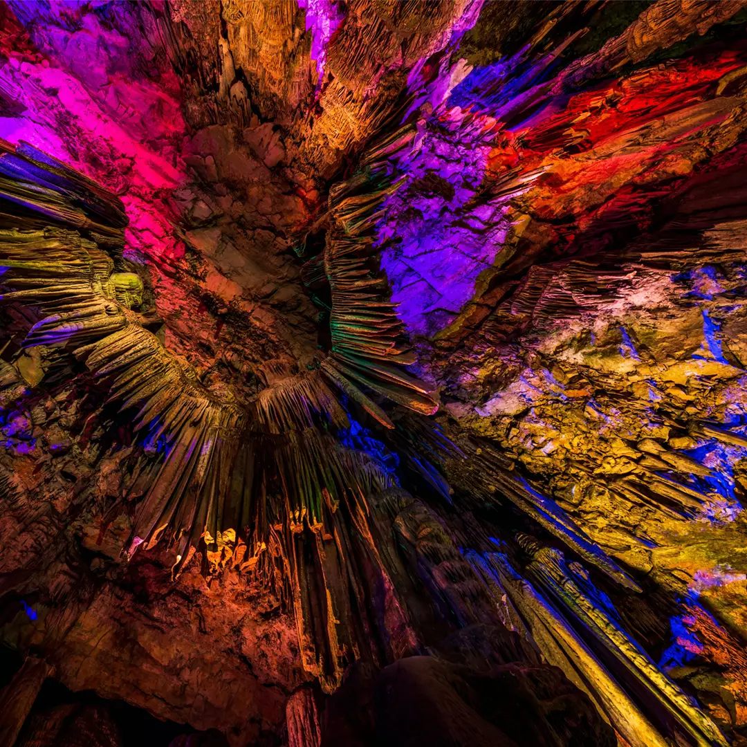 Colorful lights illuminate cave stalactites and rock formations, highlighting textures and creating a vibrant, multicolored display on the cave ceiling.