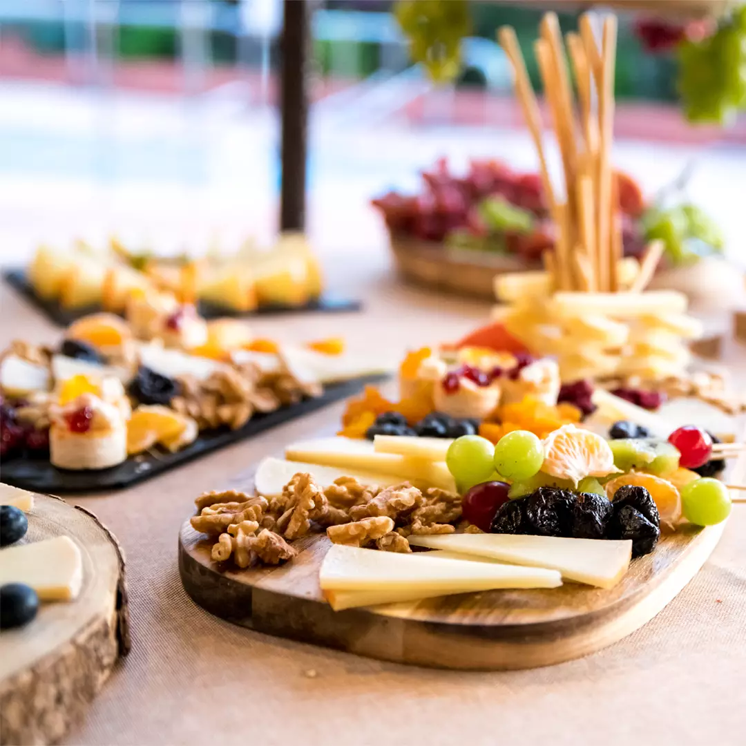 A wooden platter with assorted cheeses, walnuts, grapes, tangerines, prunes, and breadsticks, with more fruit and cheese platters in the background.