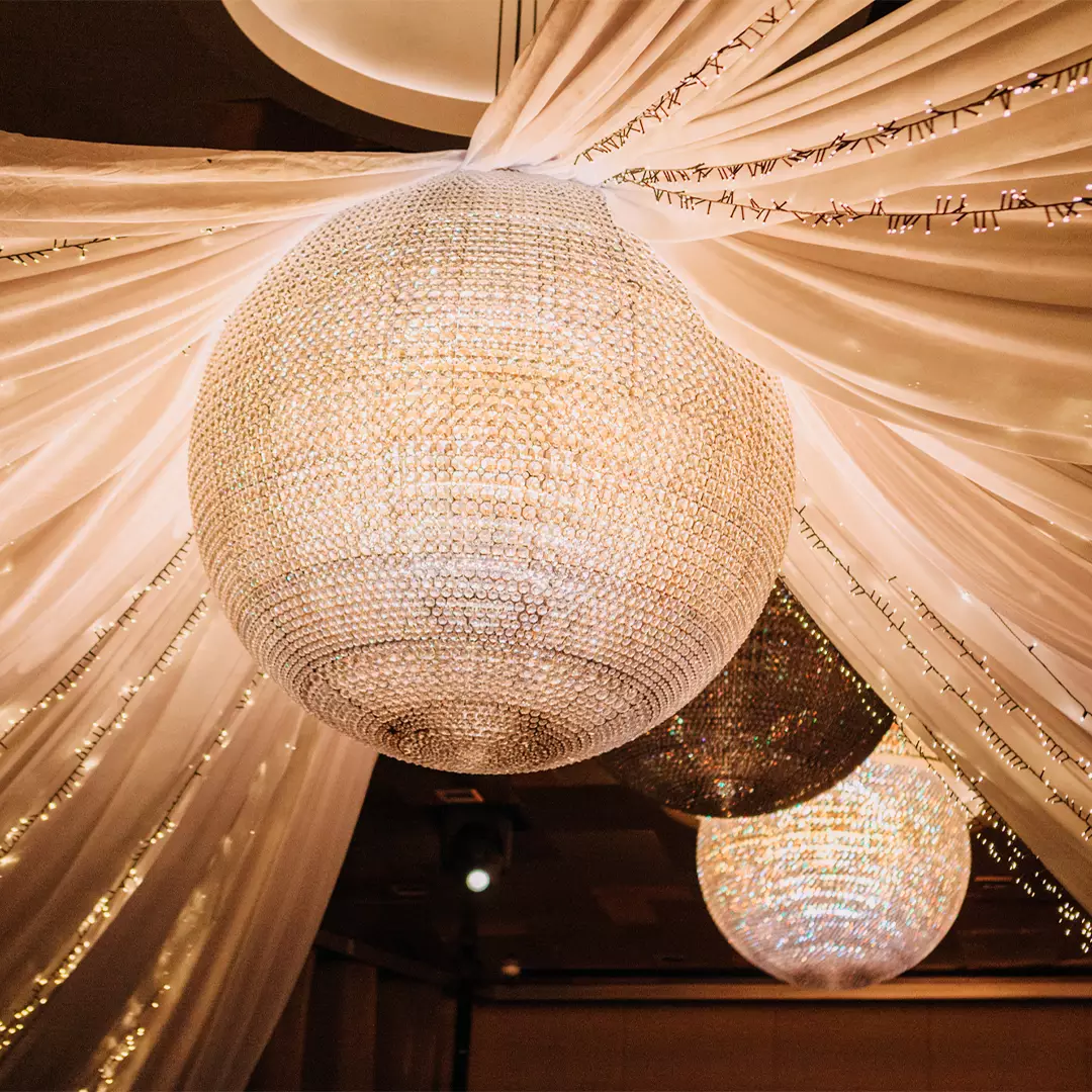 Large, spherical crystal chandeliers hang from a ceiling draped with white fabric and string lights, creating a decorative, illuminated setting.