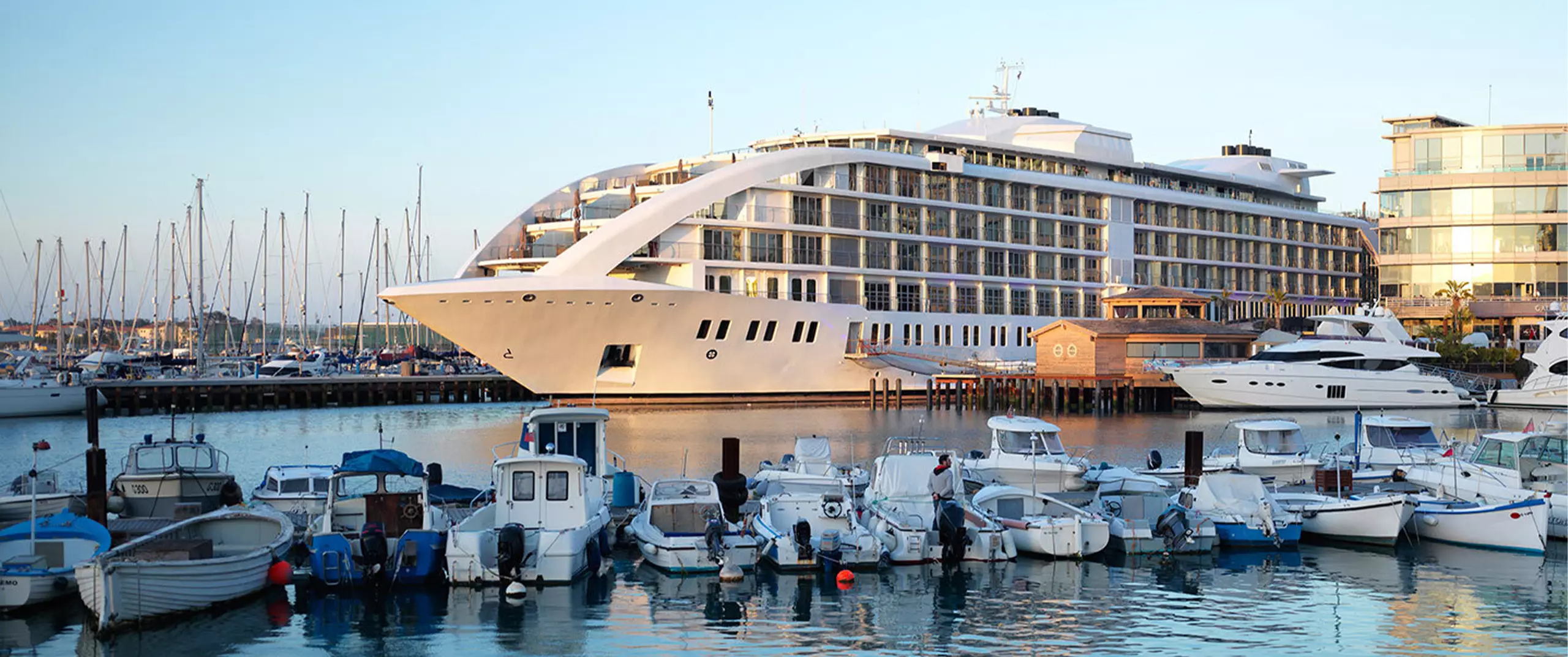 A large, modern yacht-like building docked at a marina, surrounded by smaller boats and calm water, with other yachts and a glass-fronted building nearby.