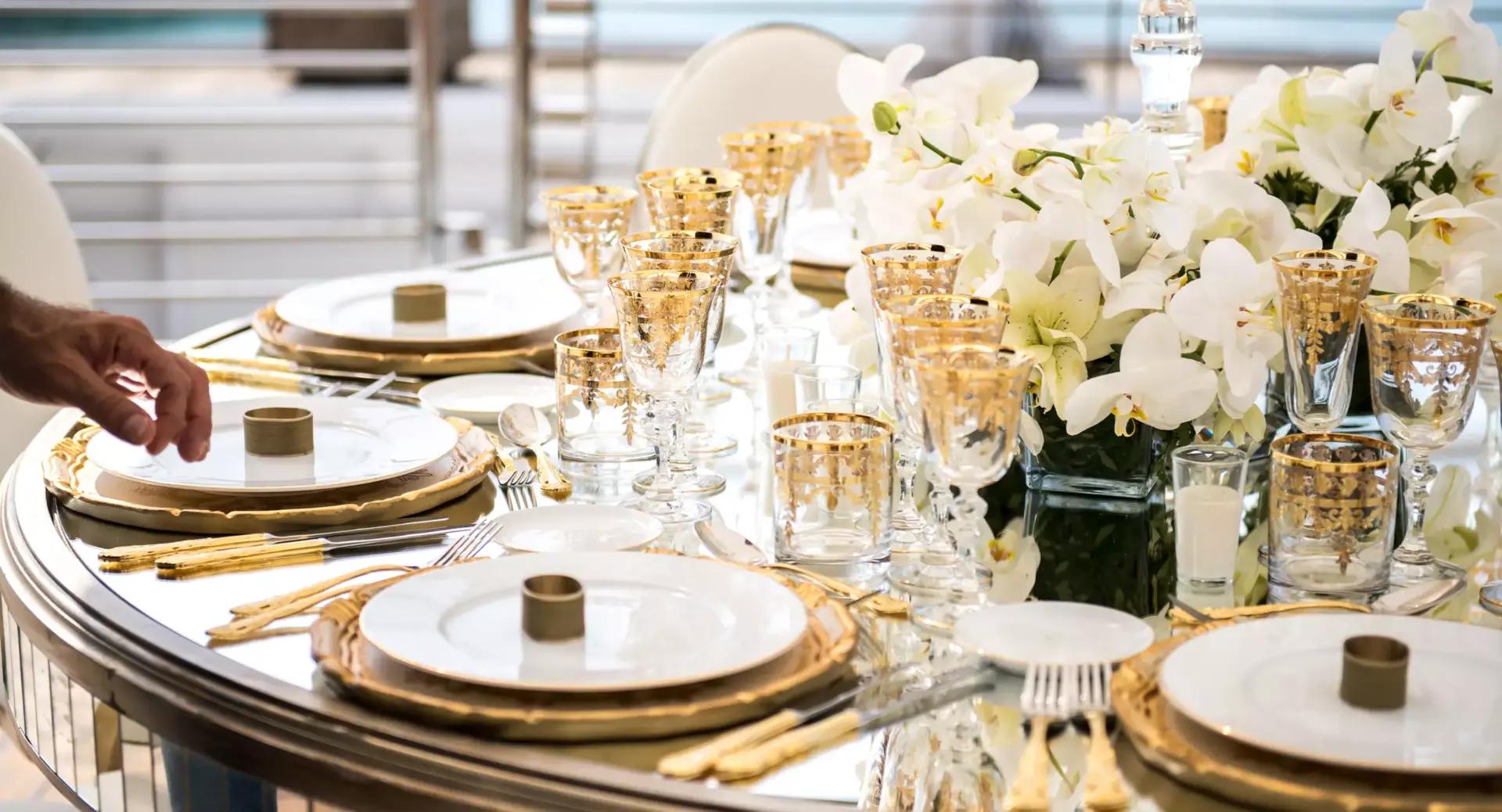 A formal dining table is set with white plates, gold-rimmed glasses, gold cutlery, and a centerpiece of white flowers; a person’s hand arranges a plate.